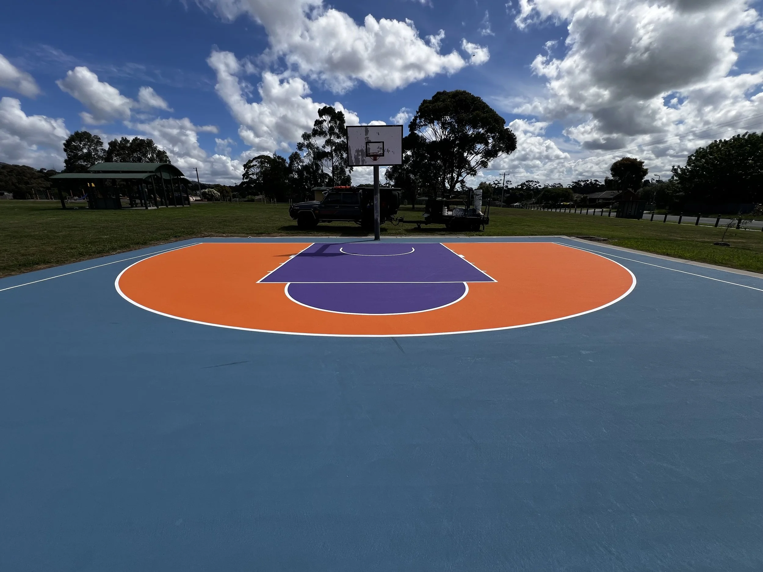 Colorful outdoor basketball court with blue, orange, and purple markings under a partly cloudy sky, with trees and vehicles in the background.