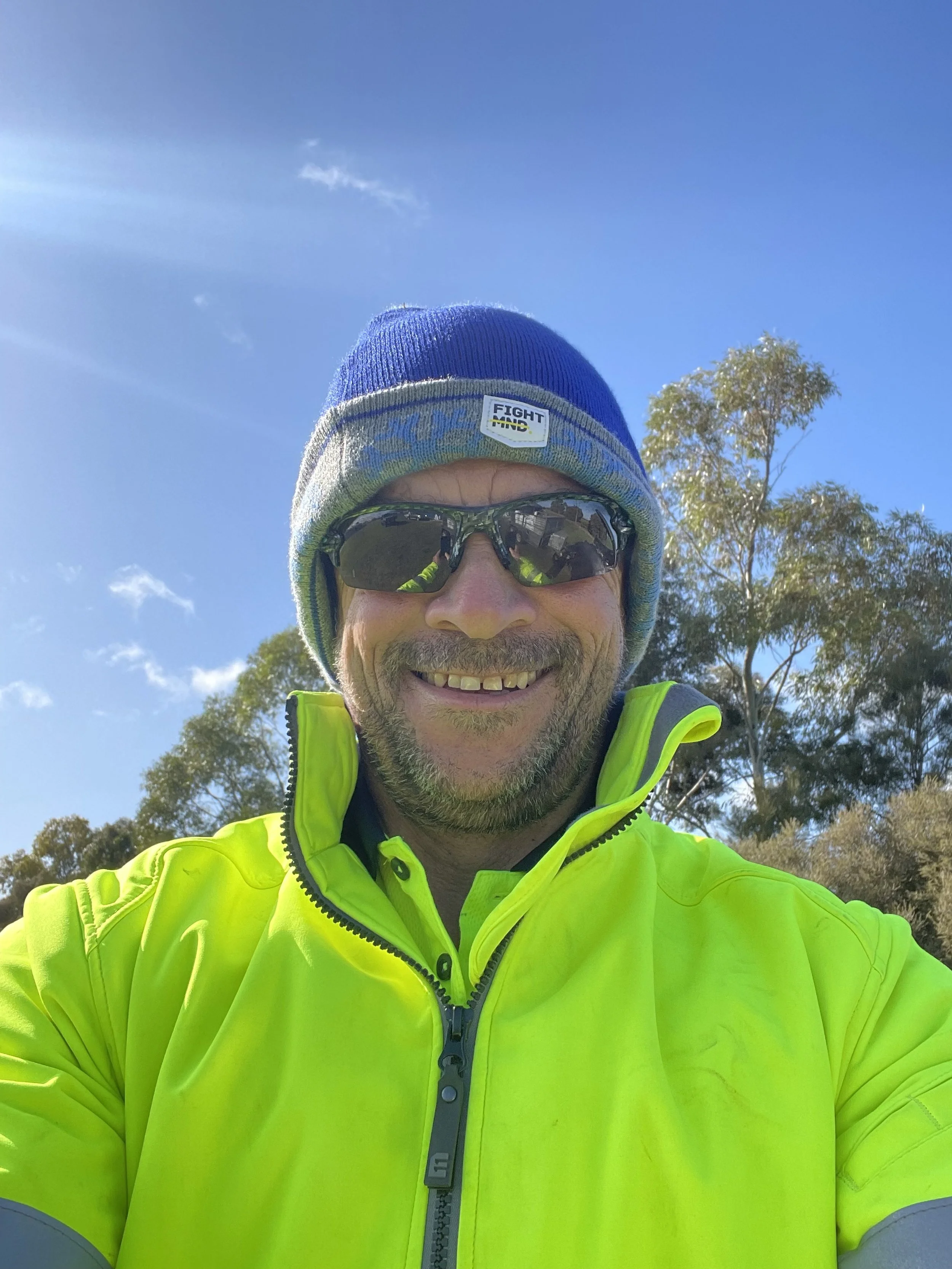 A man outdoors wearing a blue and gray beanie with a "FIGHT MNC" patch, sunglasses, and a bright yellow high-visibility jacket, smiling at the camera with trees and a blue sky in the background.