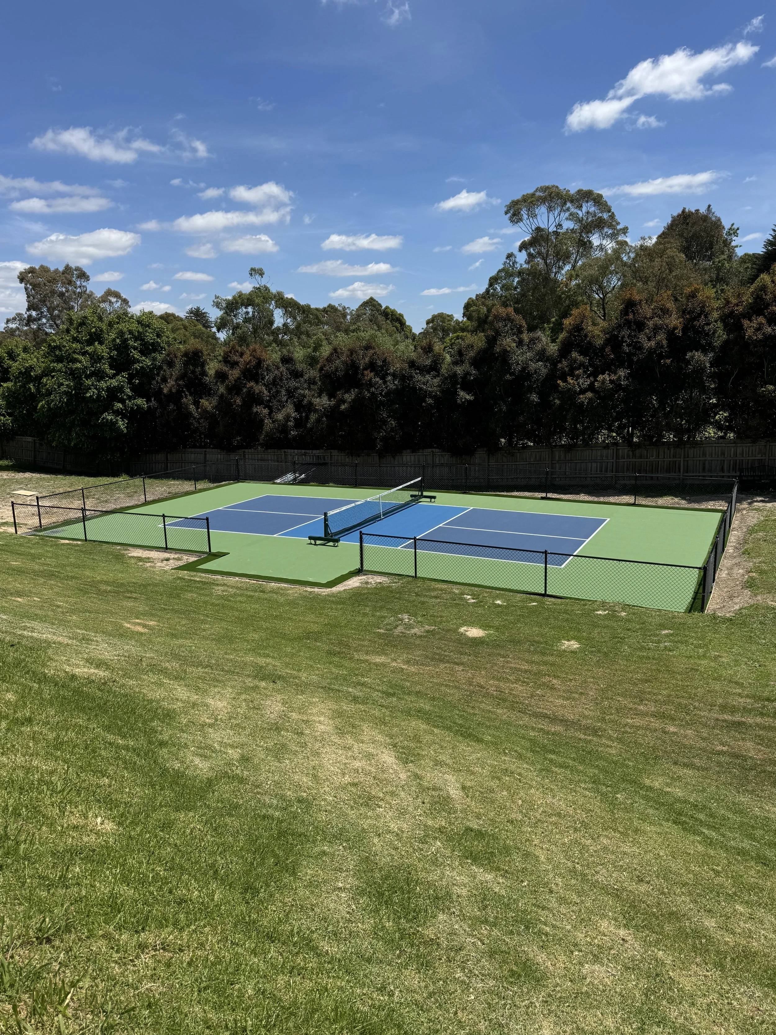 An outdoor tennis court with blue playing surface, surrounded by a green area and a black chain-link fence, under a partly cloudy sky with trees in the background.