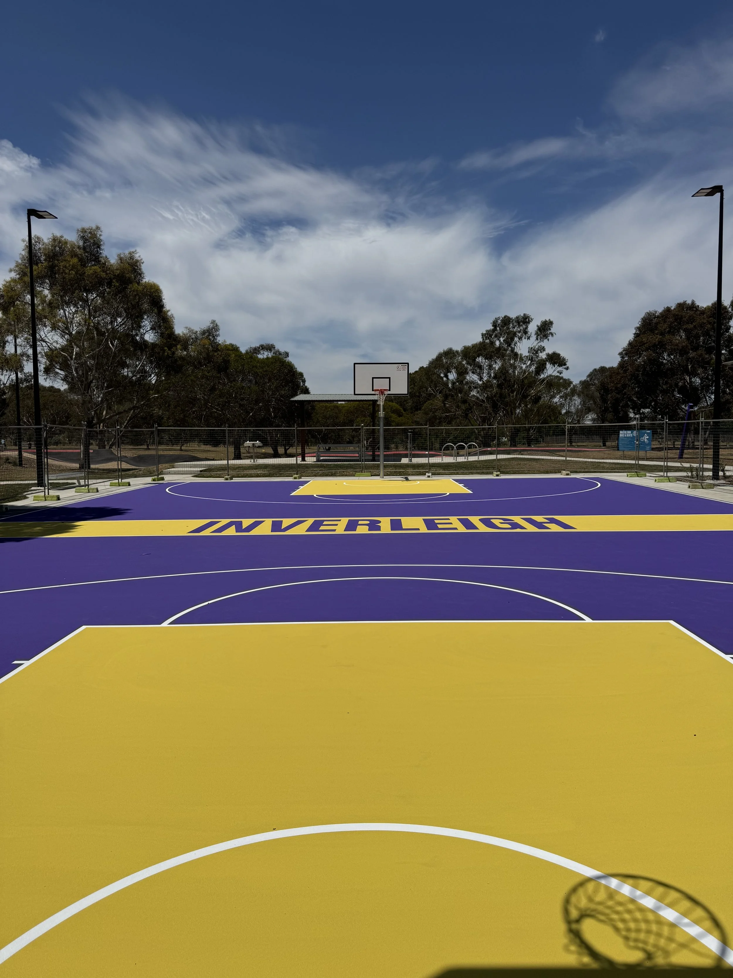 An outdoor basketball court painted in yellow and purple, with the word 'INVERLEIGH' written on the court, surrounded by trees and a fence, under a partly cloudy sky.
