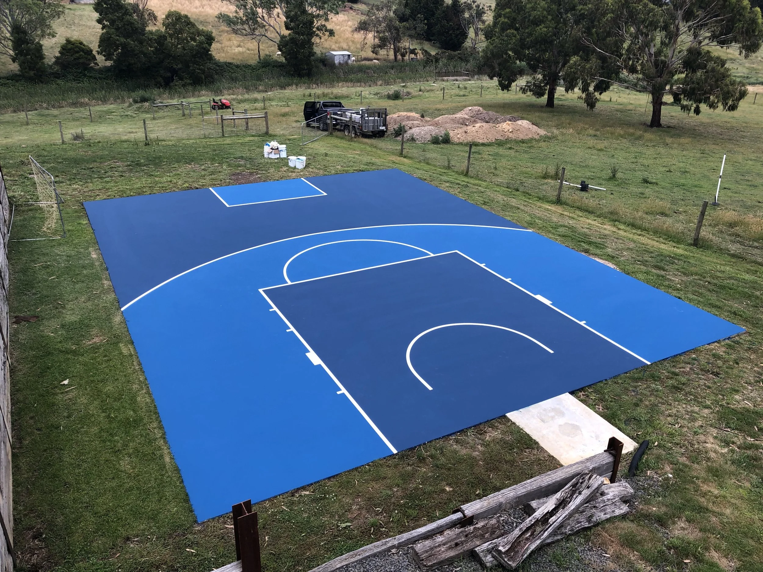 Newly painted small outdoor basketball court with blue playing surface, white lines, and a hoop, situated on a grassy area with trees and fenced yard surrounding it.
