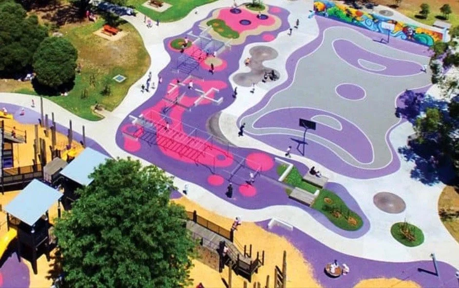 Colorful playground with various play structures, including a climbing area, slides, basketball hoops, and a large purple and gray poured-in-place rubber surface for children to play.