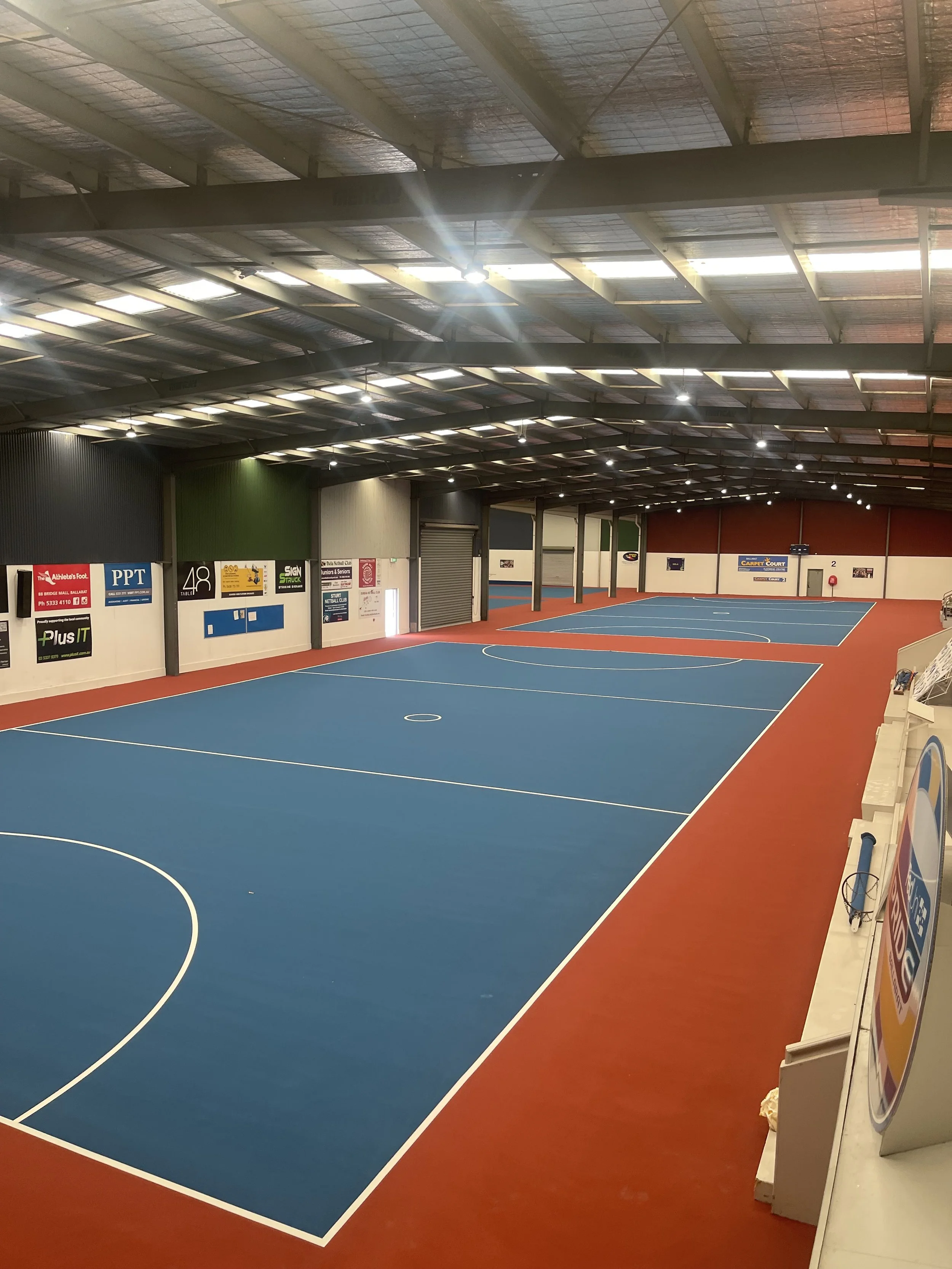 Indoor sports court with a blue floor and red border, surrounded by advertisement banners over white walls, under a metal roof with skylights.