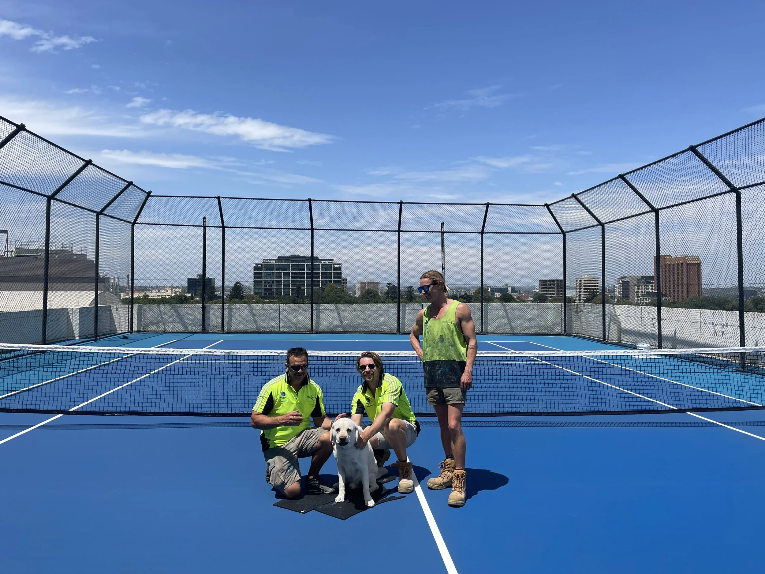 Three people and a white dog on a blue outdoor tennis or sports court with a city skyline in the background under a partly cloudy sky.