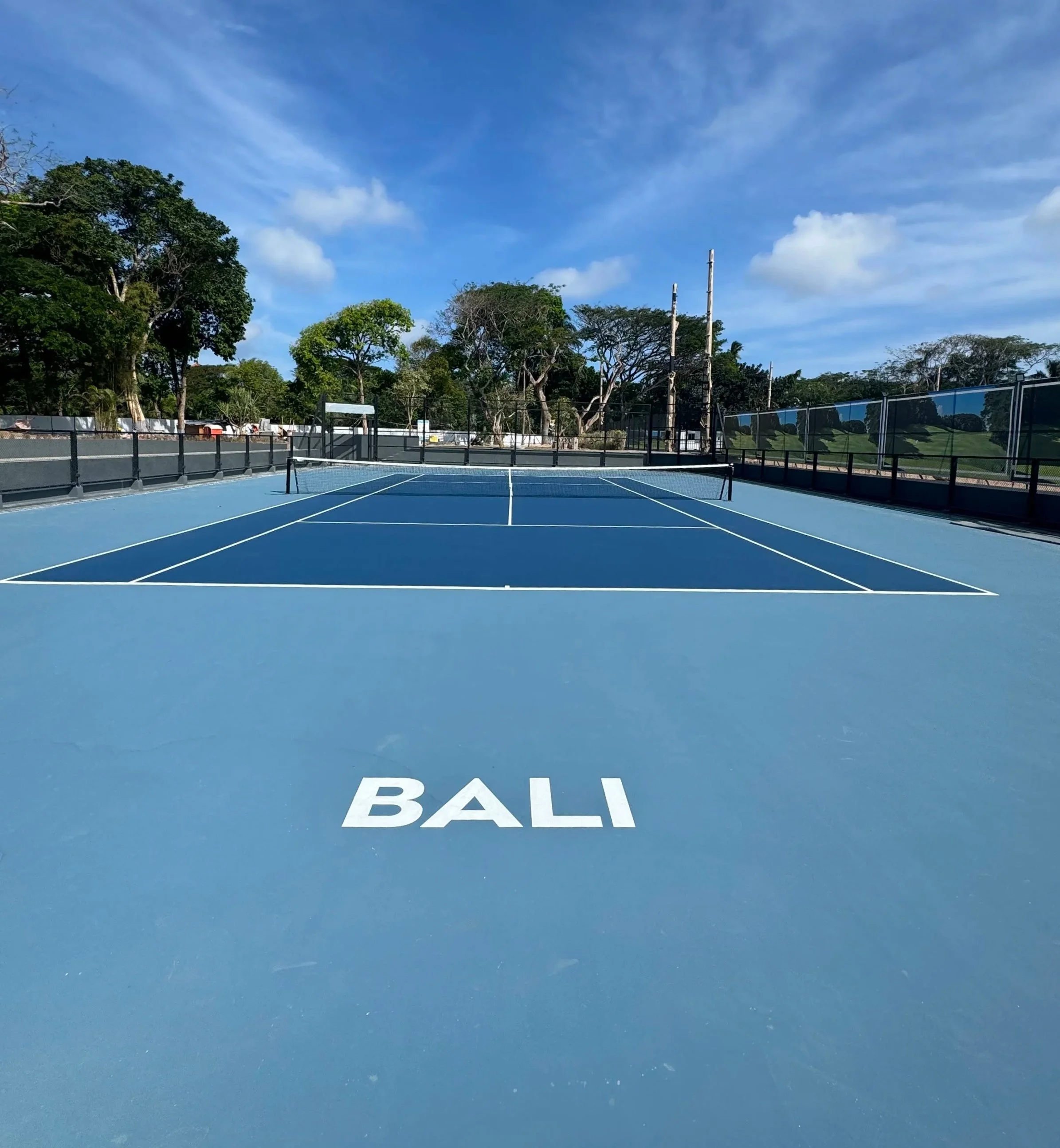 Empty outdoor tennis court with the word BALI written on the surface, surrounded by trees and a cloudy blue sky.
