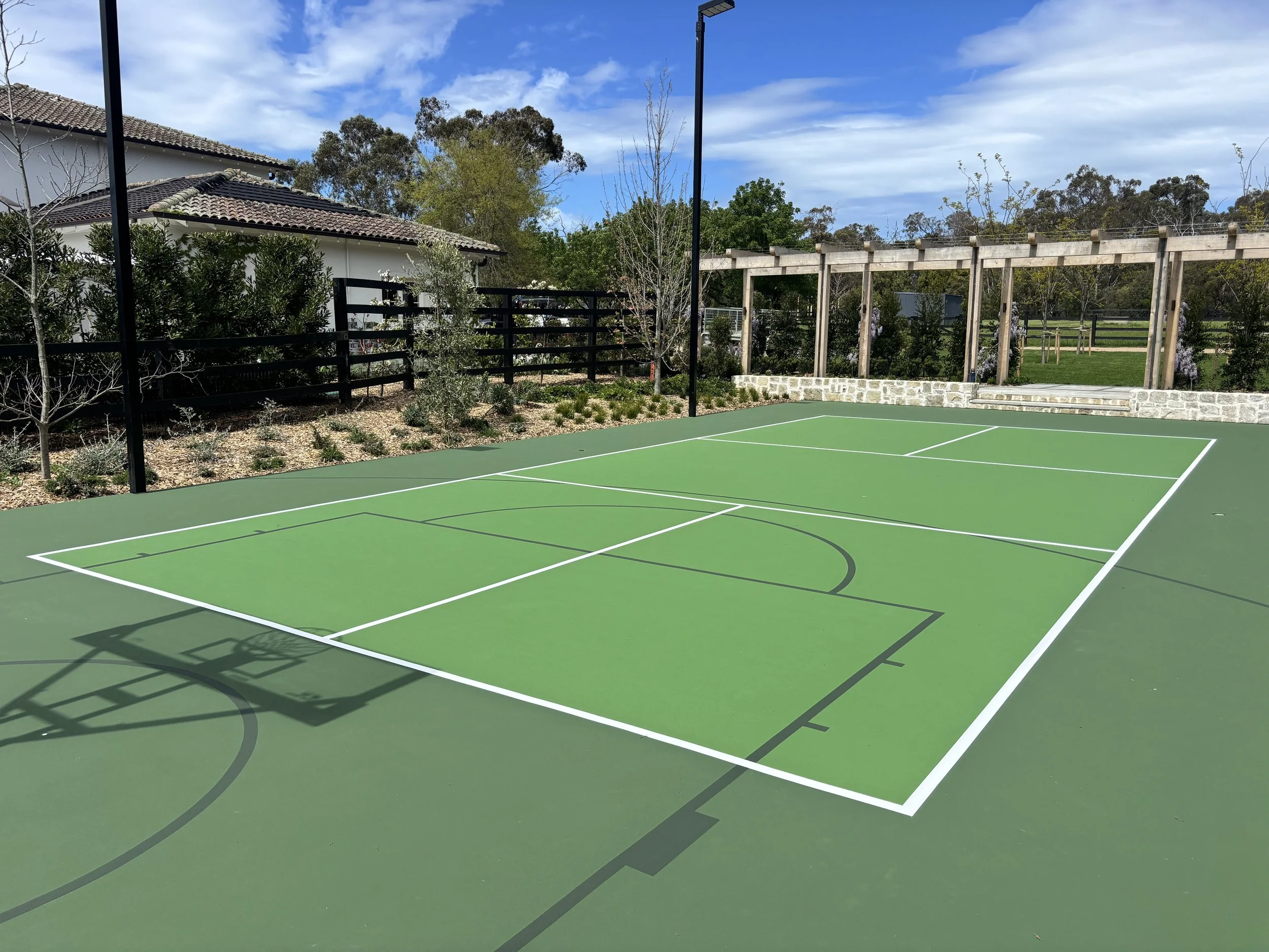 An outdoor tennis or basketball court with a green surface, white and black lines, surrounded by trees, a black fence, and a clear blue sky.