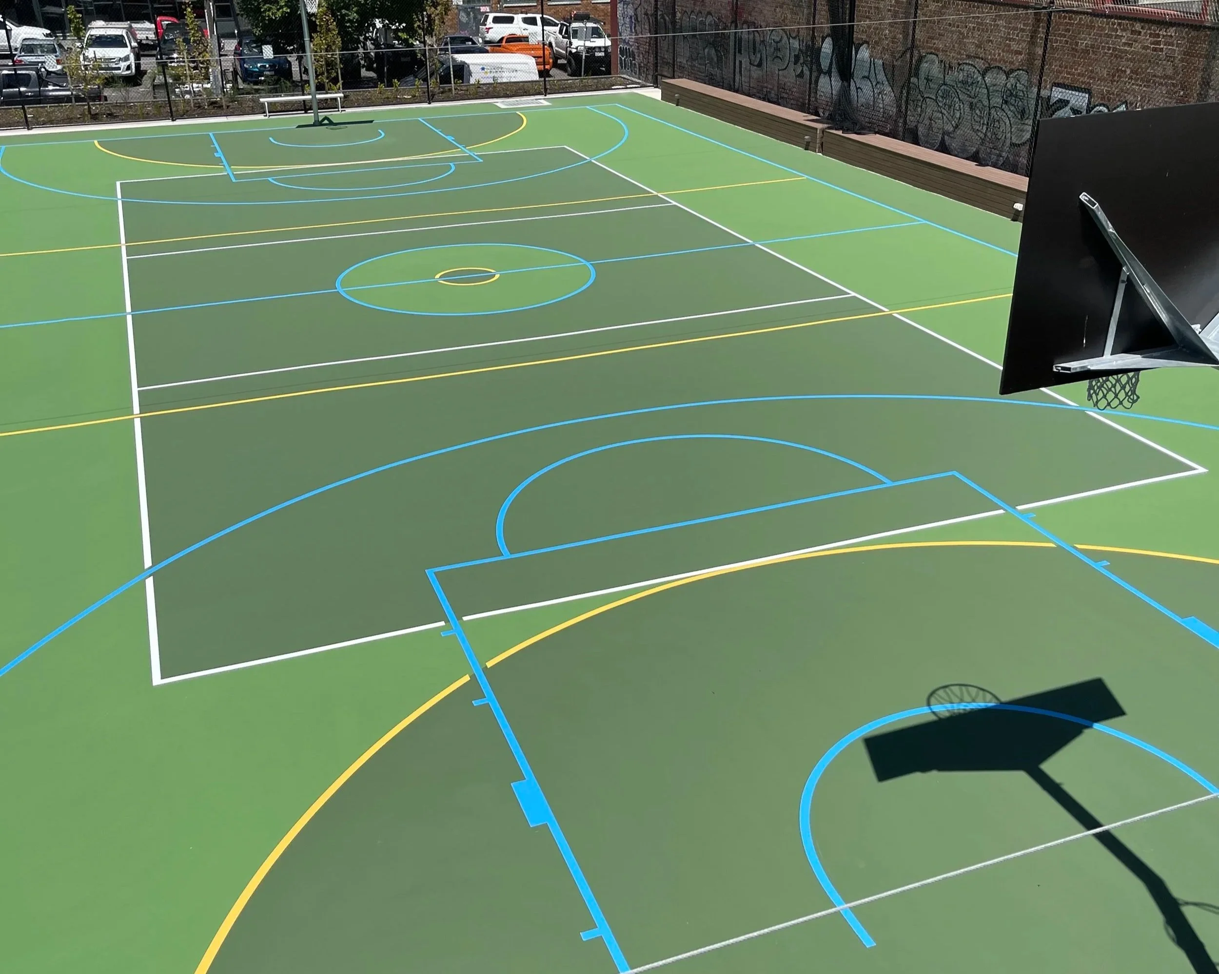 Outdoor basketball court with green surface, marked with white, blue, and yellow lines, equipped with two basketball hoops, surrounded by parked cars and a brick wall with graffiti.