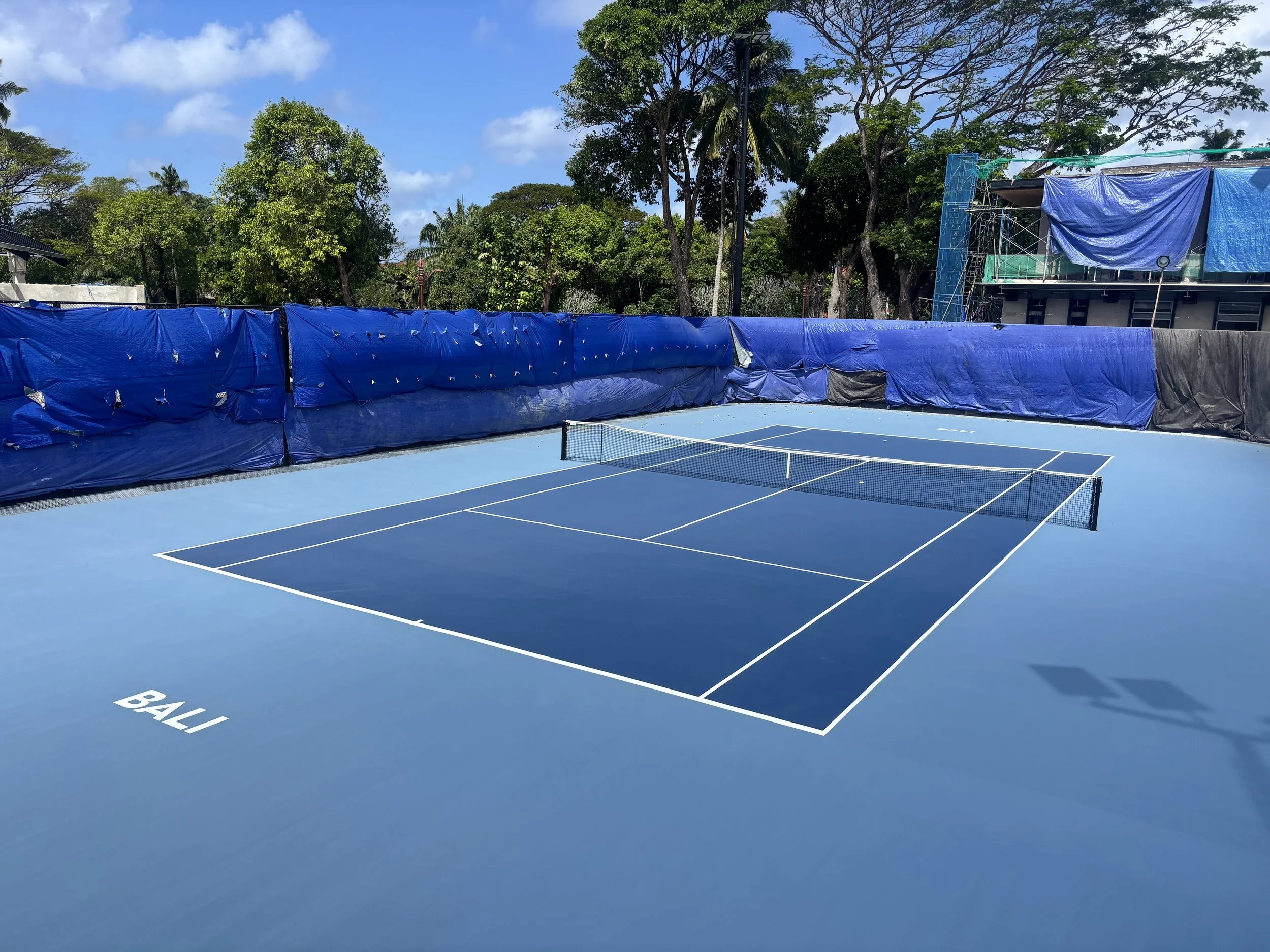 Empty outdoor tennis court with blue surface, white lines, and a net, surrounded by blue windbreaks and green trees under a partly cloudy sky.