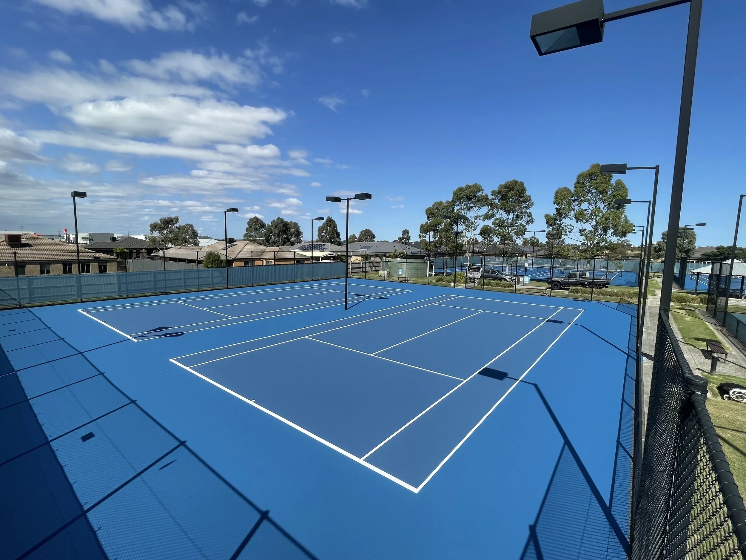 Empty blue tennis court with white lines, surrounded by a black fence, with trees, houses, parked cars, and a blue sky with clouds in the background.