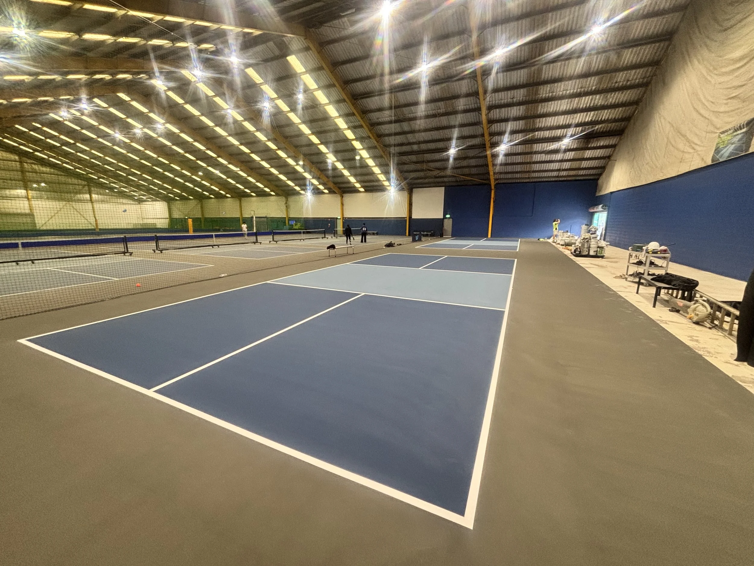 Indoor tennis courts with blue and gray surfaces, set up for practice or play, inside a large industrial-style building with a high, curved ceiling and bright overhead lighting.