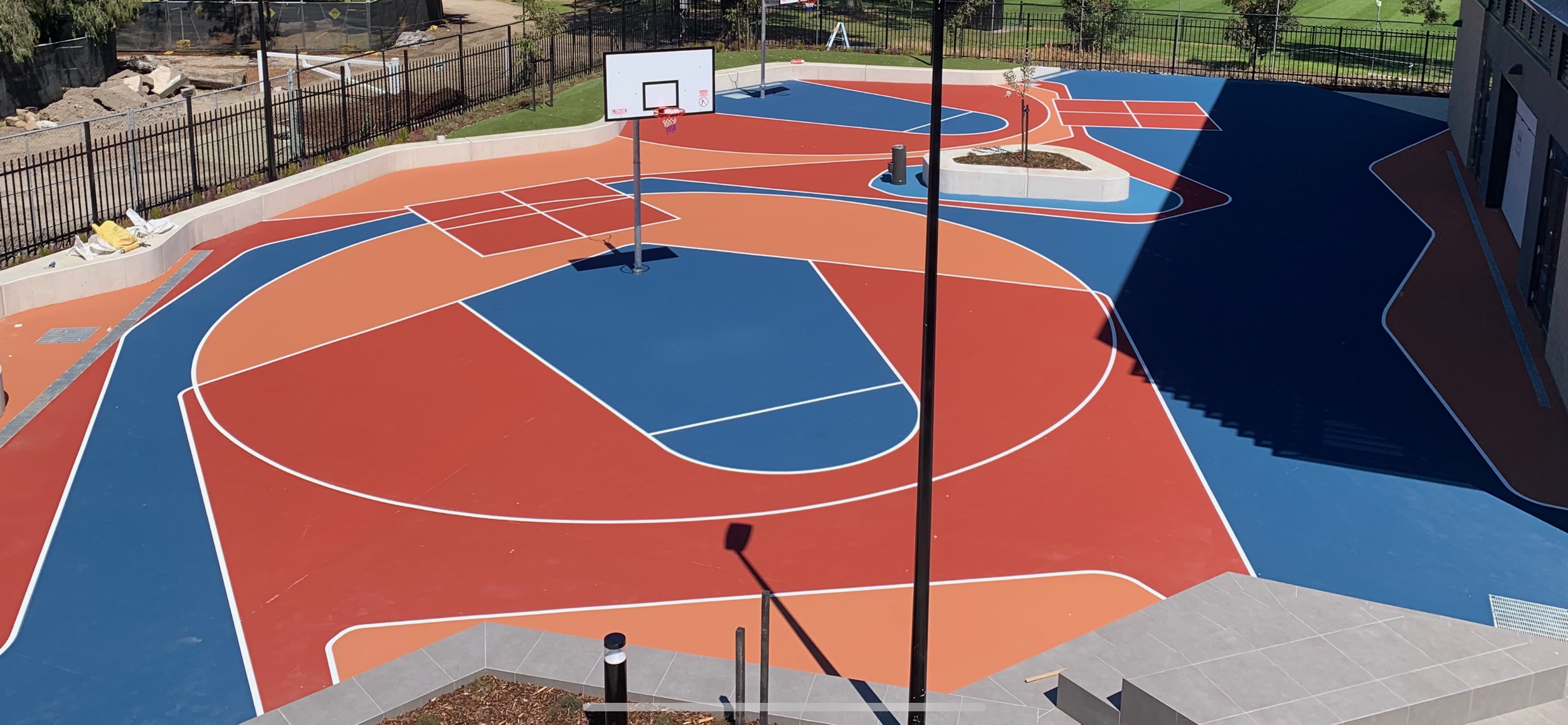 Colorful outdoor basketball court with orange and blue sections, white lines, basketball hoops, surrounded by a black fence and some trees, with stairs and a building nearby.