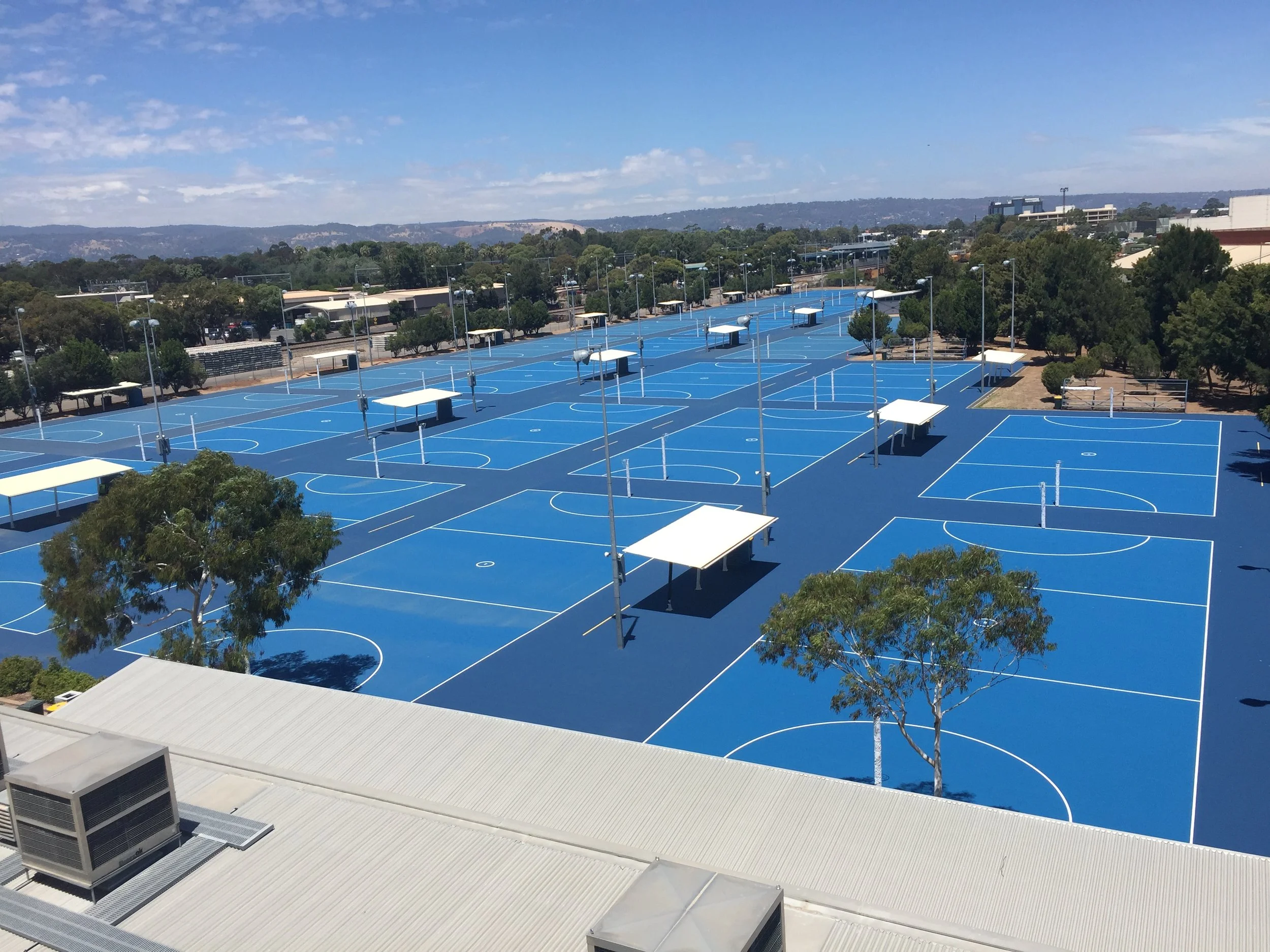 Multiple blue basketball courts with white lines, covered seating areas, and trees in a campus setting under a partly cloudy sky.