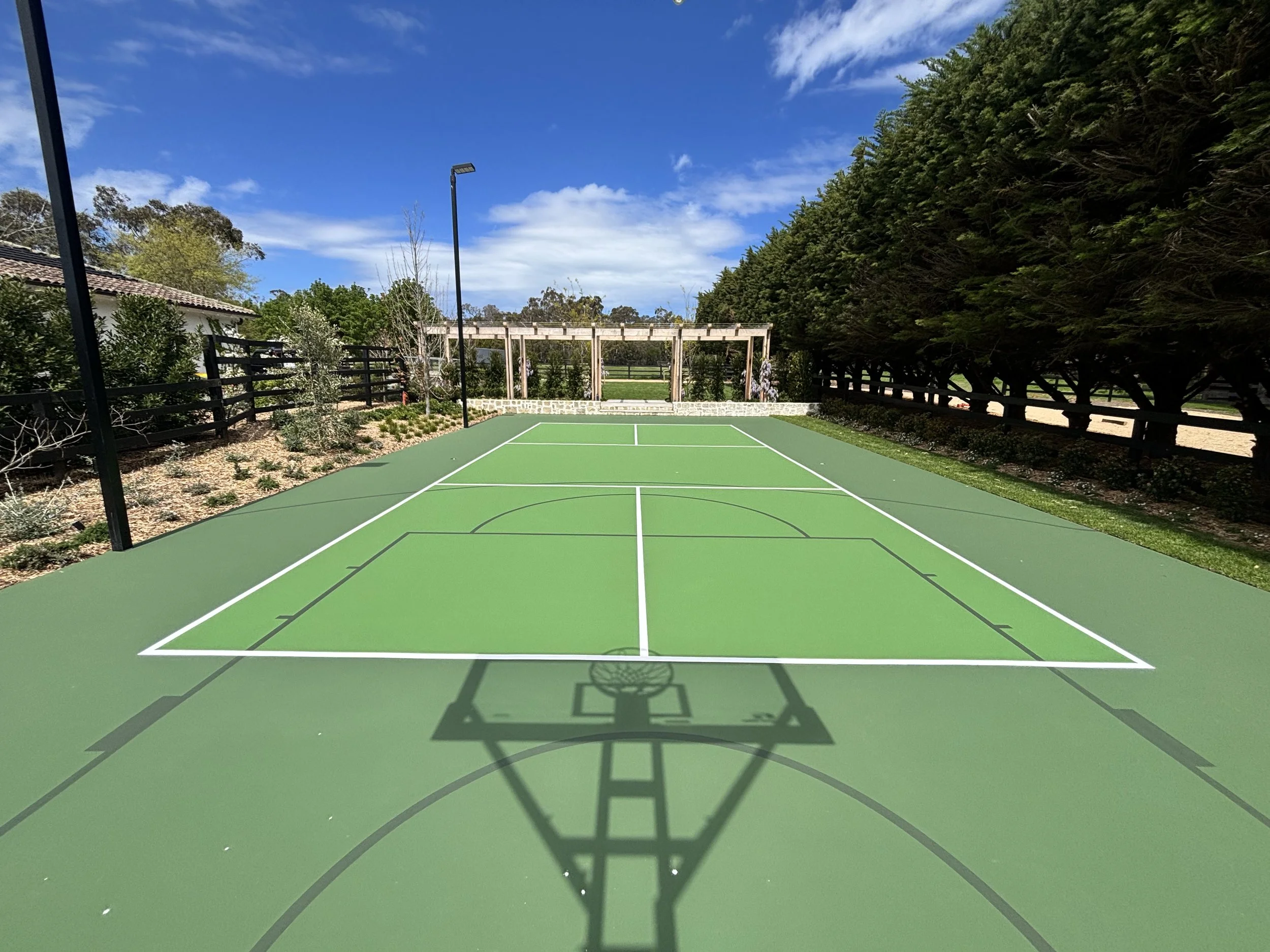 An outdoor green basketball court with white lines, surrounded by tall trees and black fences, under a partly cloudy blue sky.