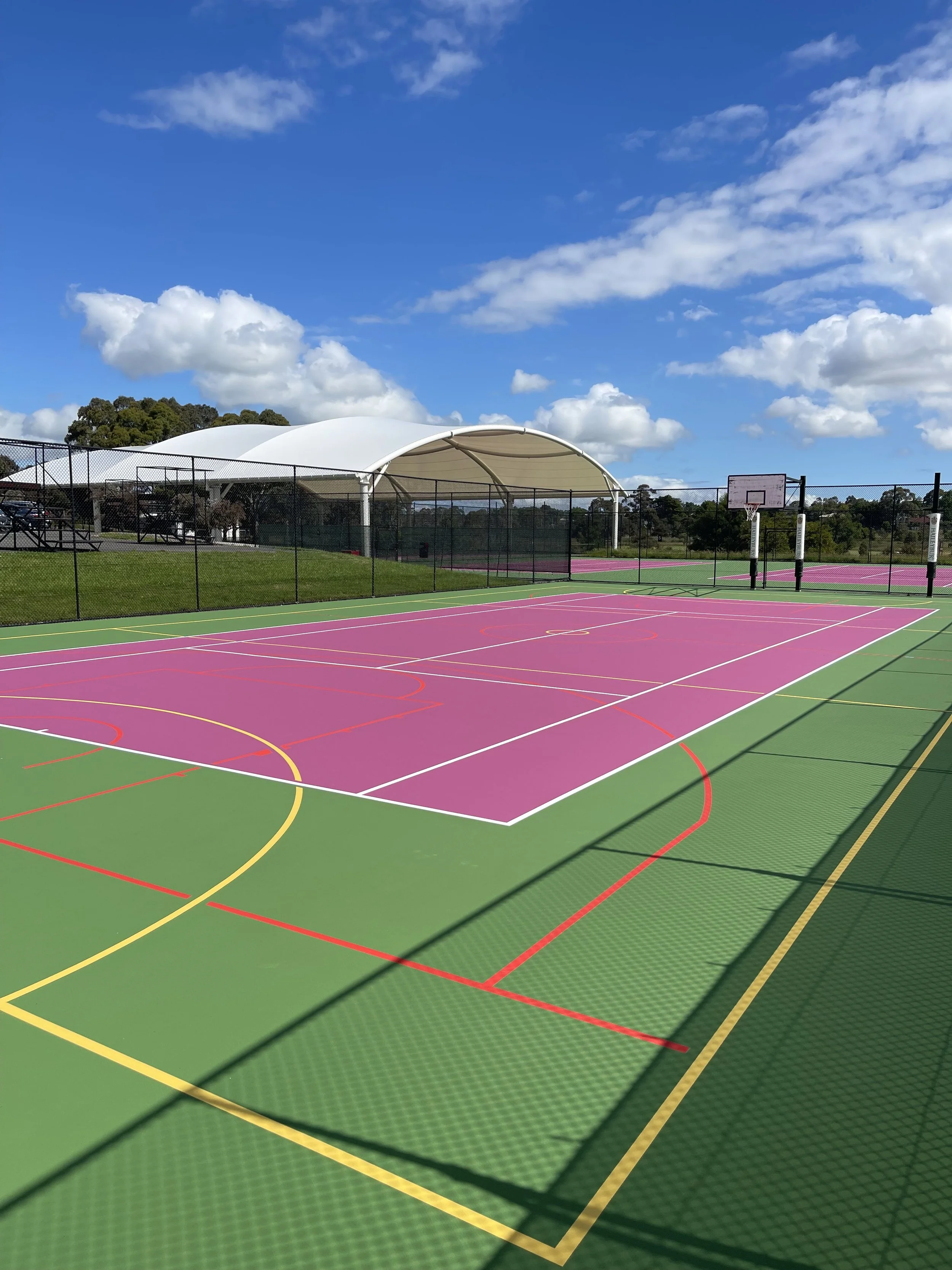 Colorful outdoor basketball court with pink playing surface and white, yellow, red lines, surrounded by a black fence, with a white canopy shelter and trees in the background under a blue sky with scattered clouds.