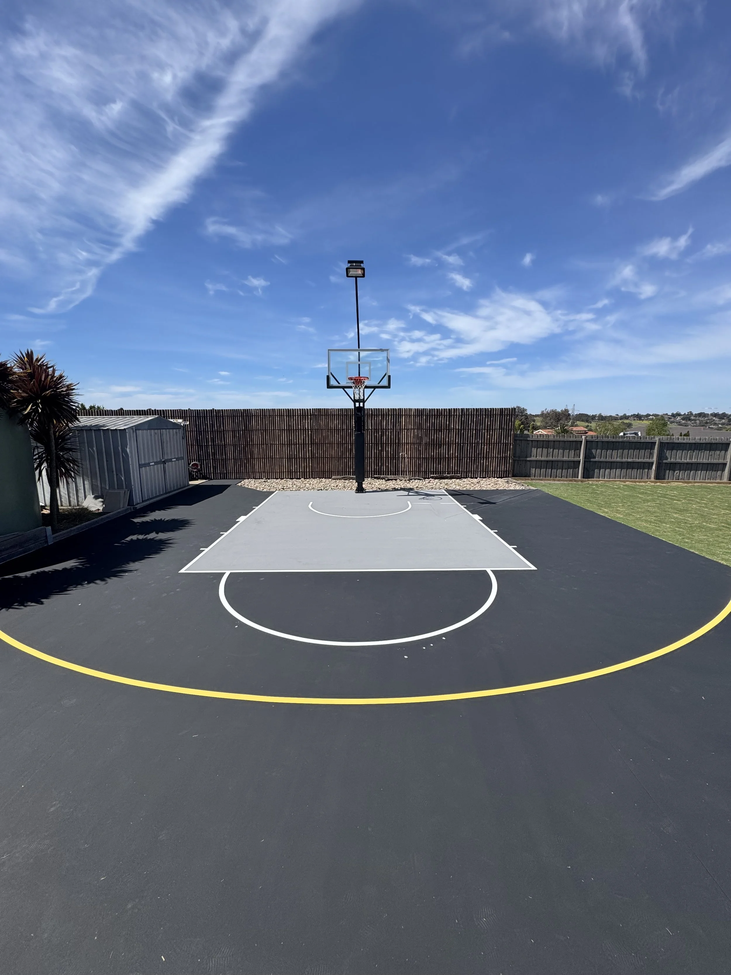An outdoor basketball court with a backboard, hoop, and painted white and yellow lines, under a partly cloudy sky.