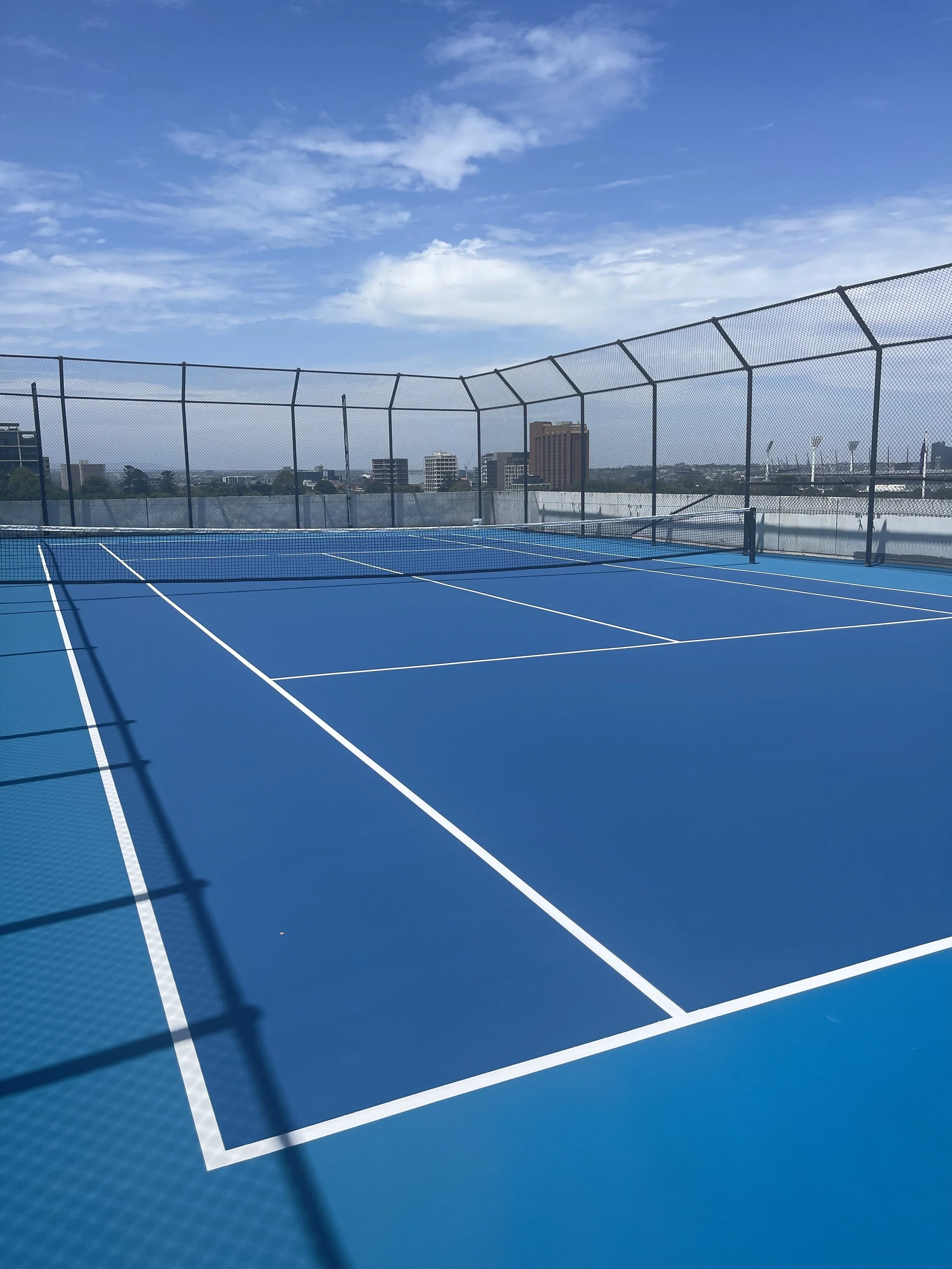 Empty blue tennis court with white lines, surrounded by a black fence, with a city skyline and blue sky with clouds in the background.