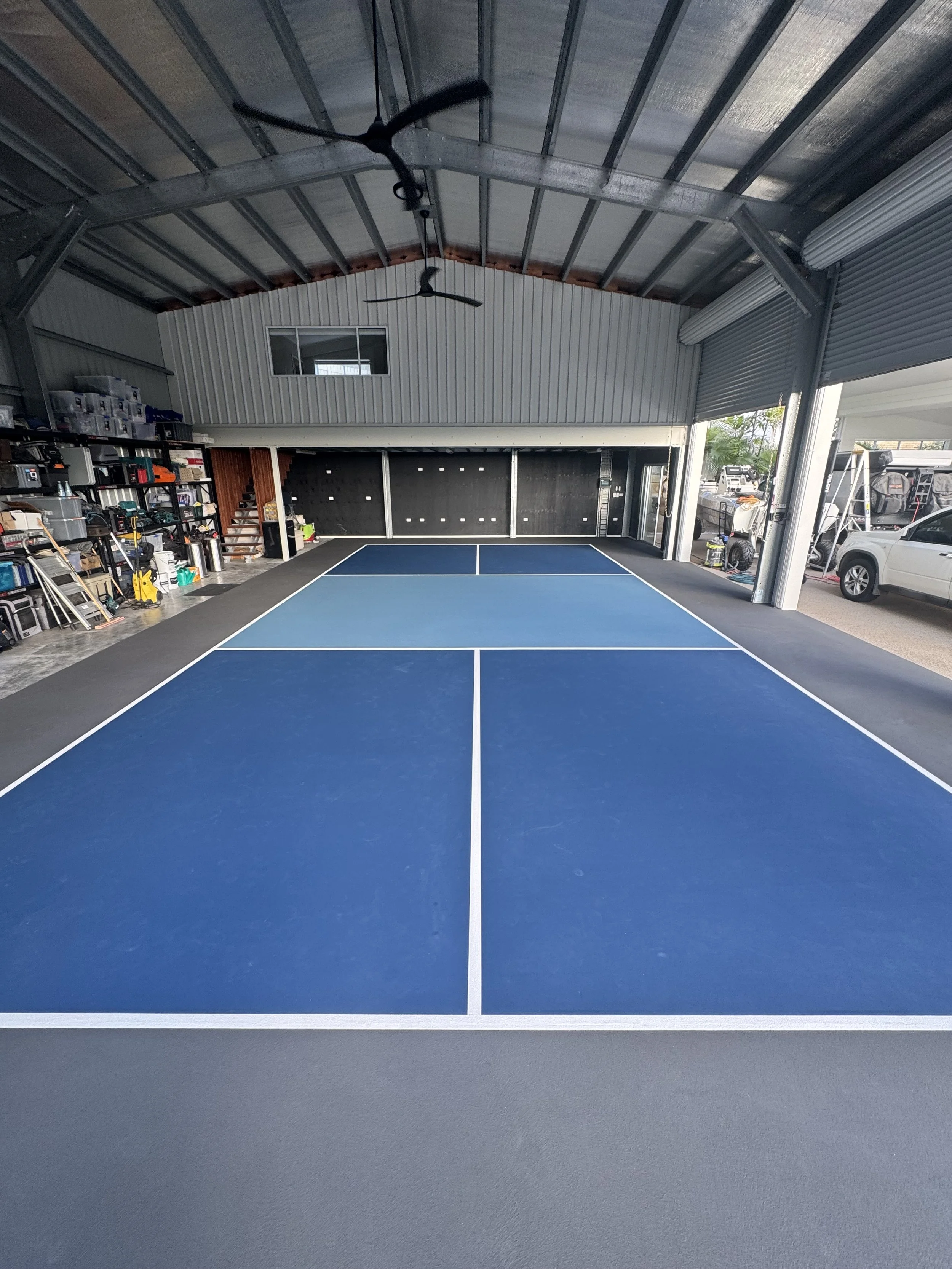 Indoor garage with a blue tennis or pickleball court, exposed metal roof, and parked vehicles outside. Storage shelves and equipment on the left side, with a small window above the court.