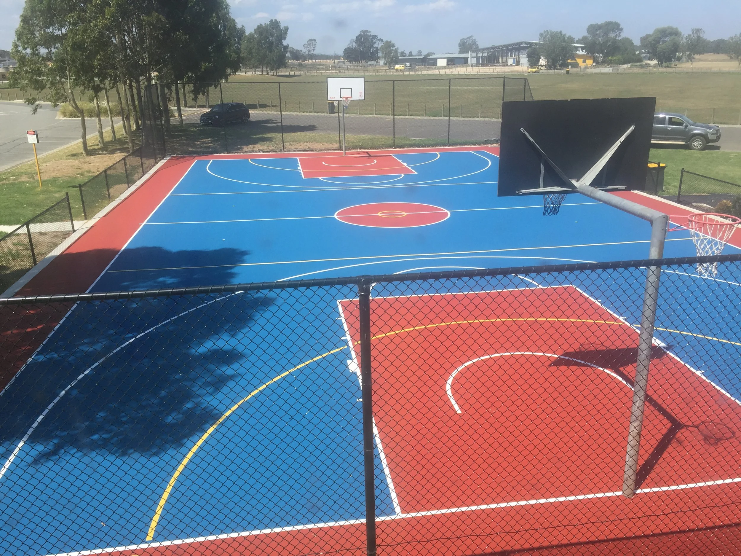 Colorful outdoor basketball court with blue, red, and yellow lines, surrounded by a black chain-link fence, with trees, cars, and a building in the background.