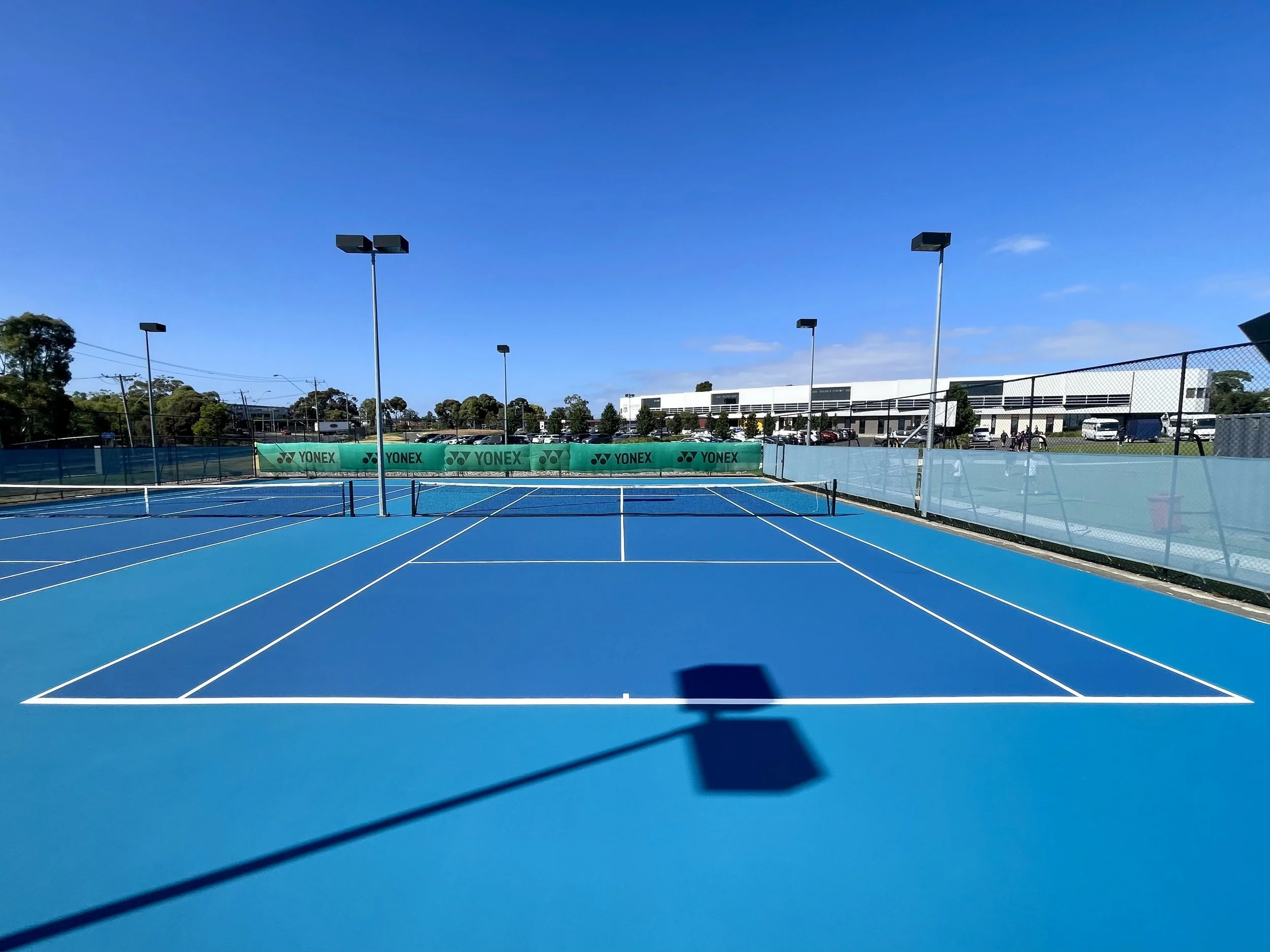 Empty blue tennis court outdoors with white lines, surrounded by a fence, with several tall light poles and trees in the background under a clear blue sky.