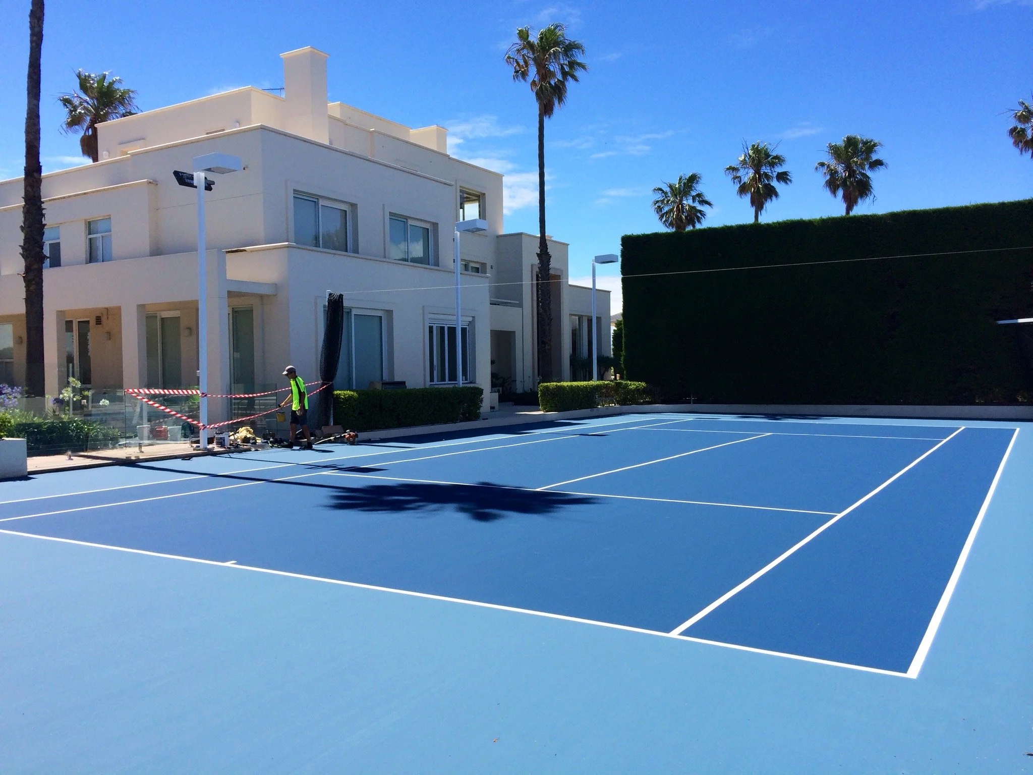 A newly painted blue tennis court with white lines at a modern residential area. There are some workers and equipment near the courts, with palm trees and a large white apartment building in the background under a blue sky.