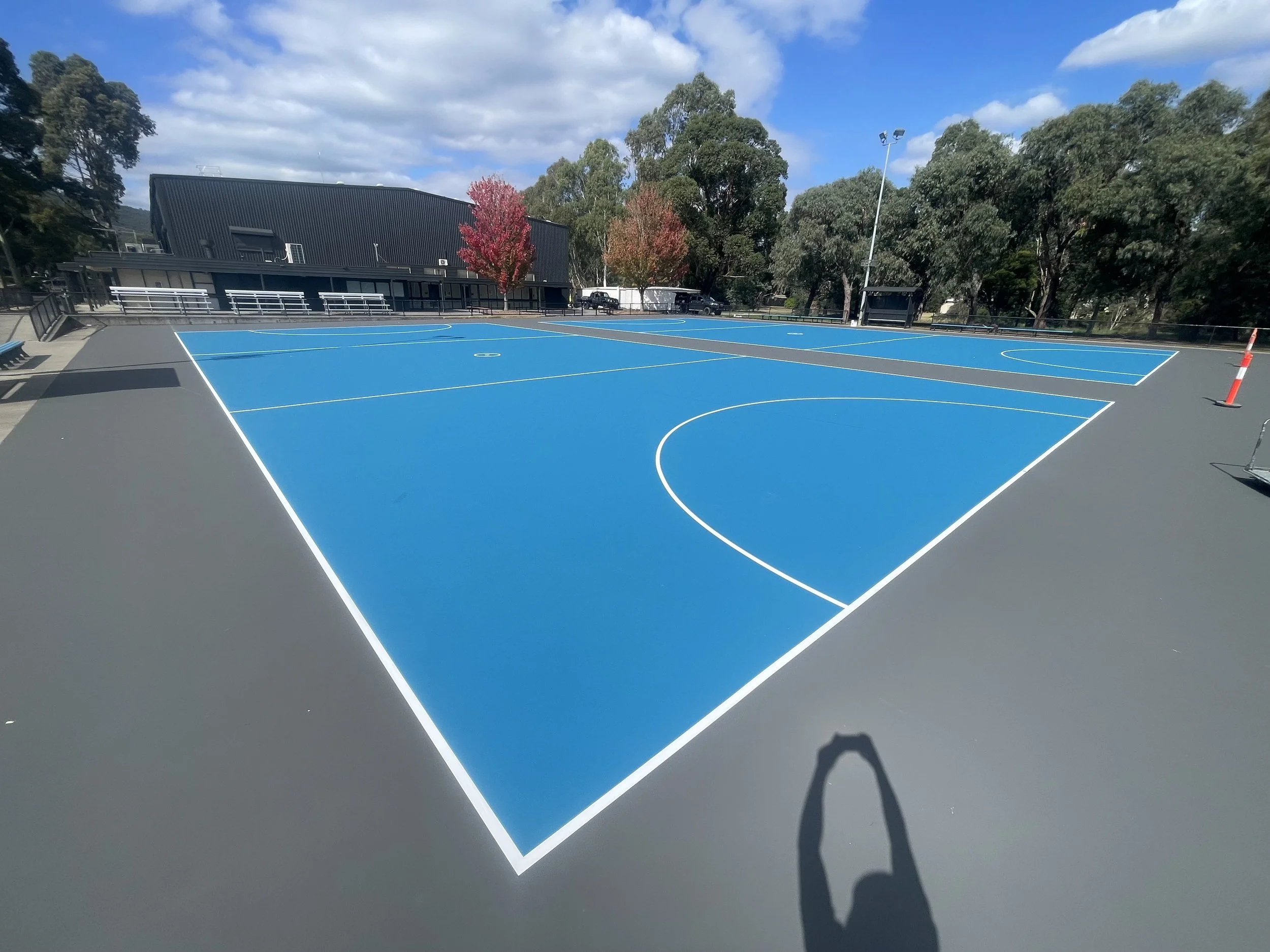 Empty outdoor basketball court with blue and gray surface, surrounded by trees and a black building, under a partly cloudy sky.