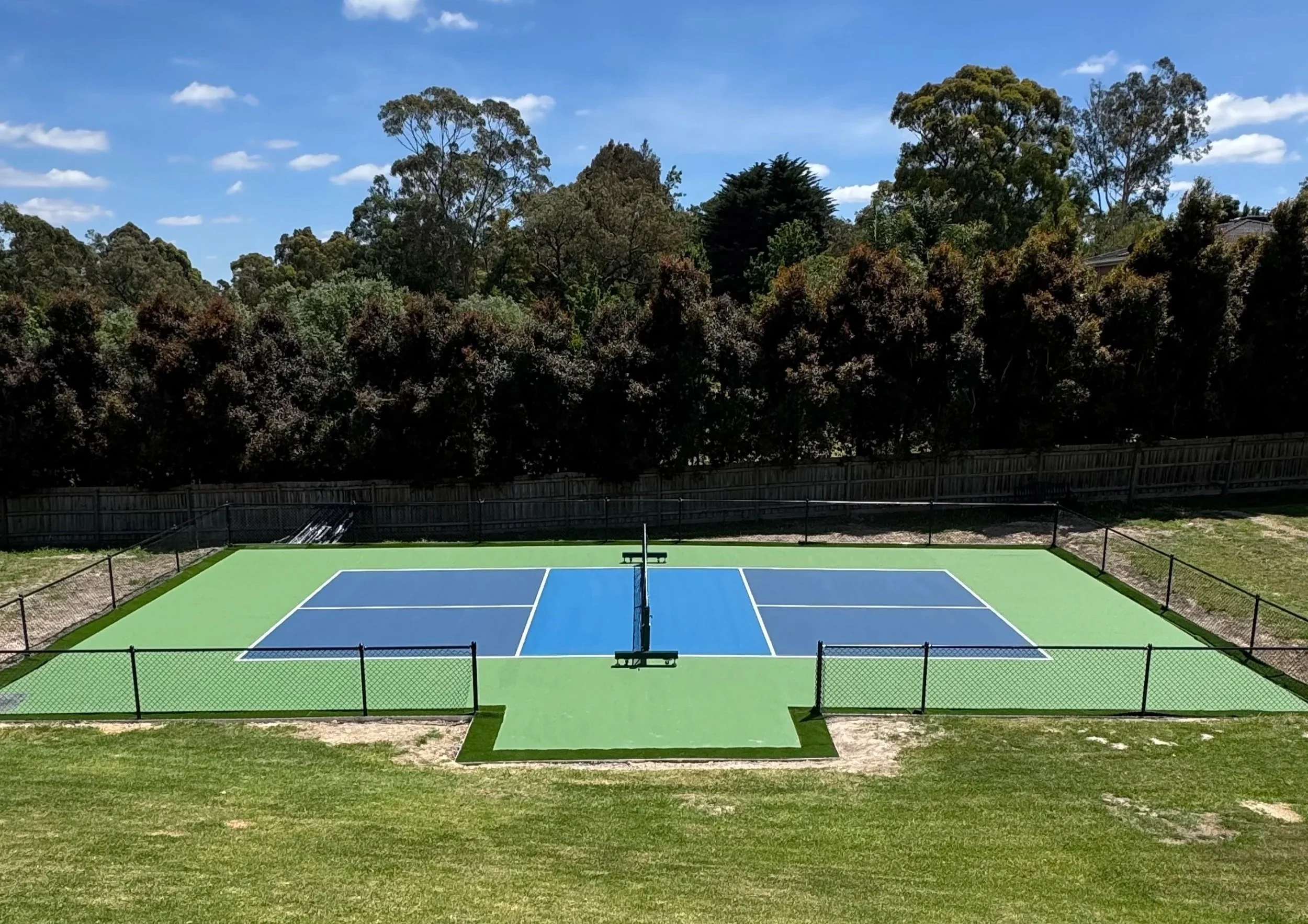 Empty tennis court with blue and green surface, surrounded by a black fence, with trees and a blue sky in the background.