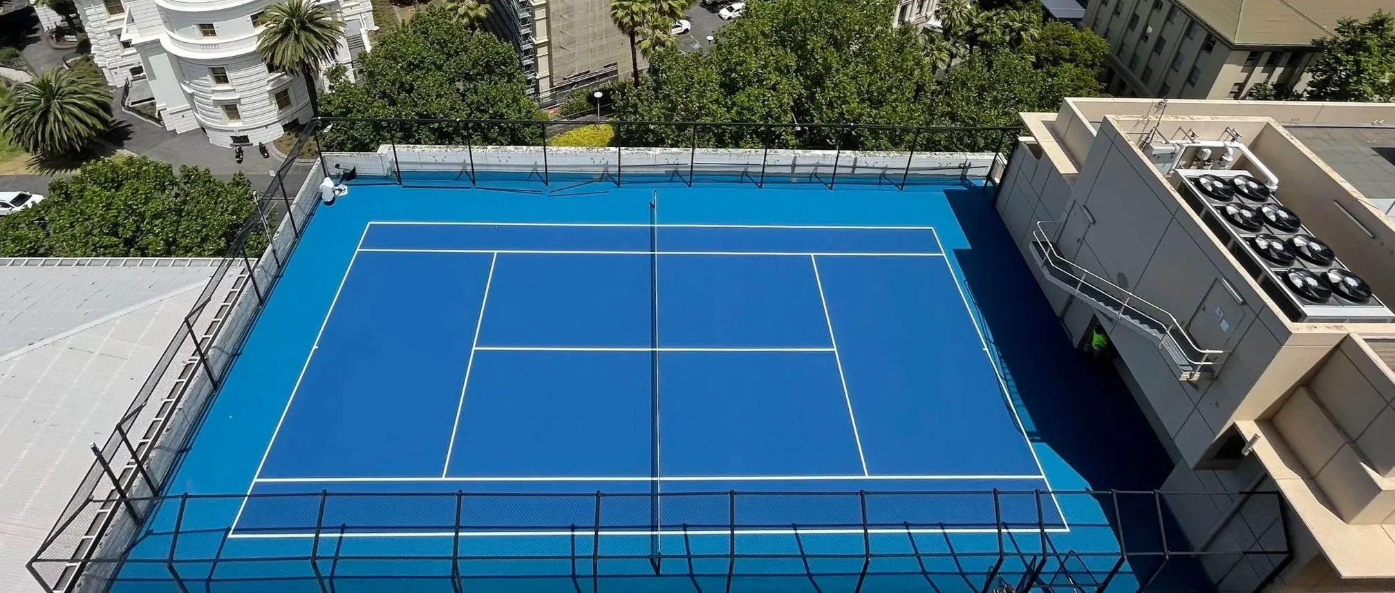 A rooftop tennis court with a blue surface, marked with white lines, surrounded by a black fence. There are buildings and trees visible around the court.
