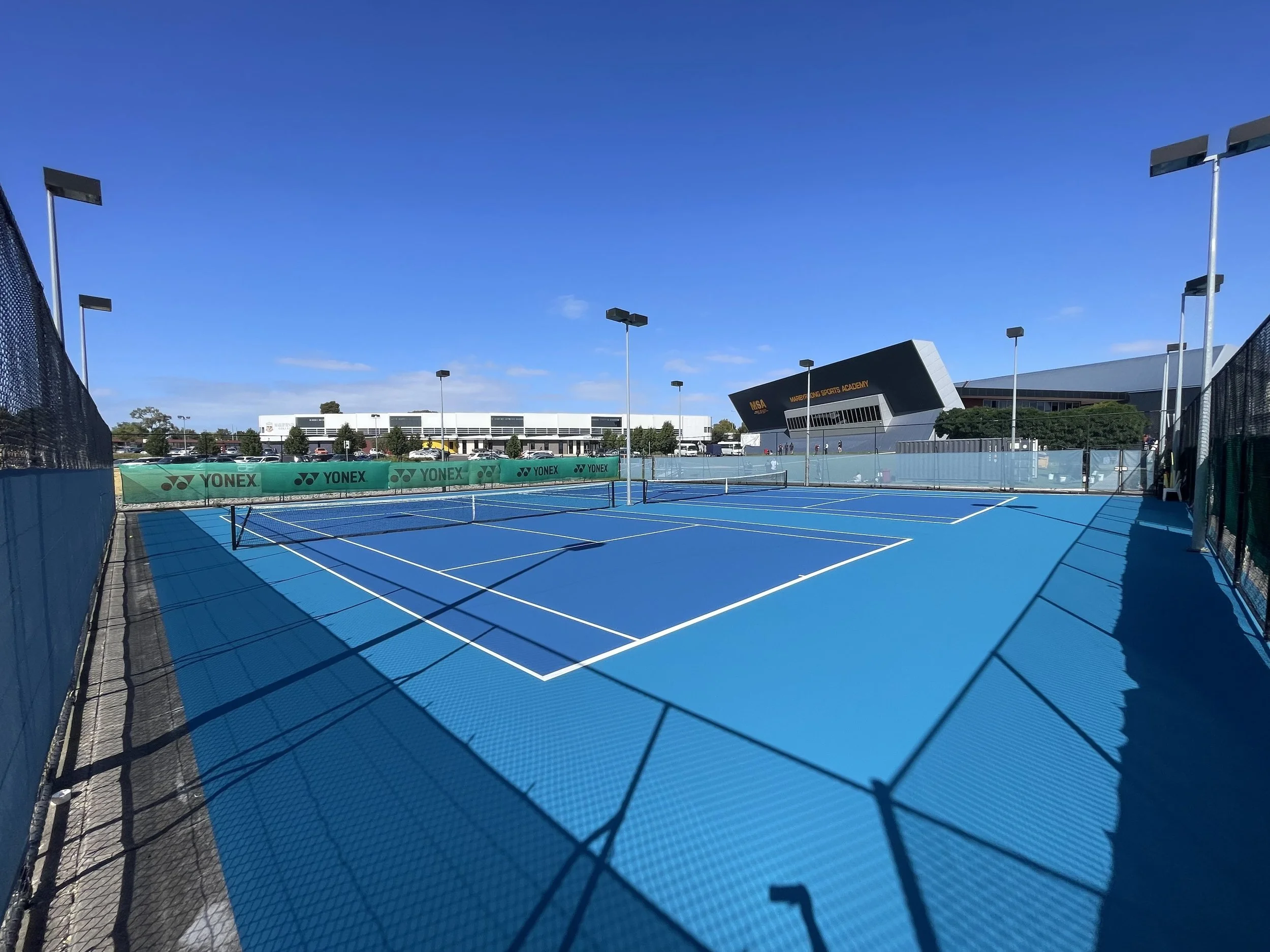 Empty blue tennis court with white lines, surrounded by a black fence with green Yonex banners, under a clear blue sky at a sports complex.