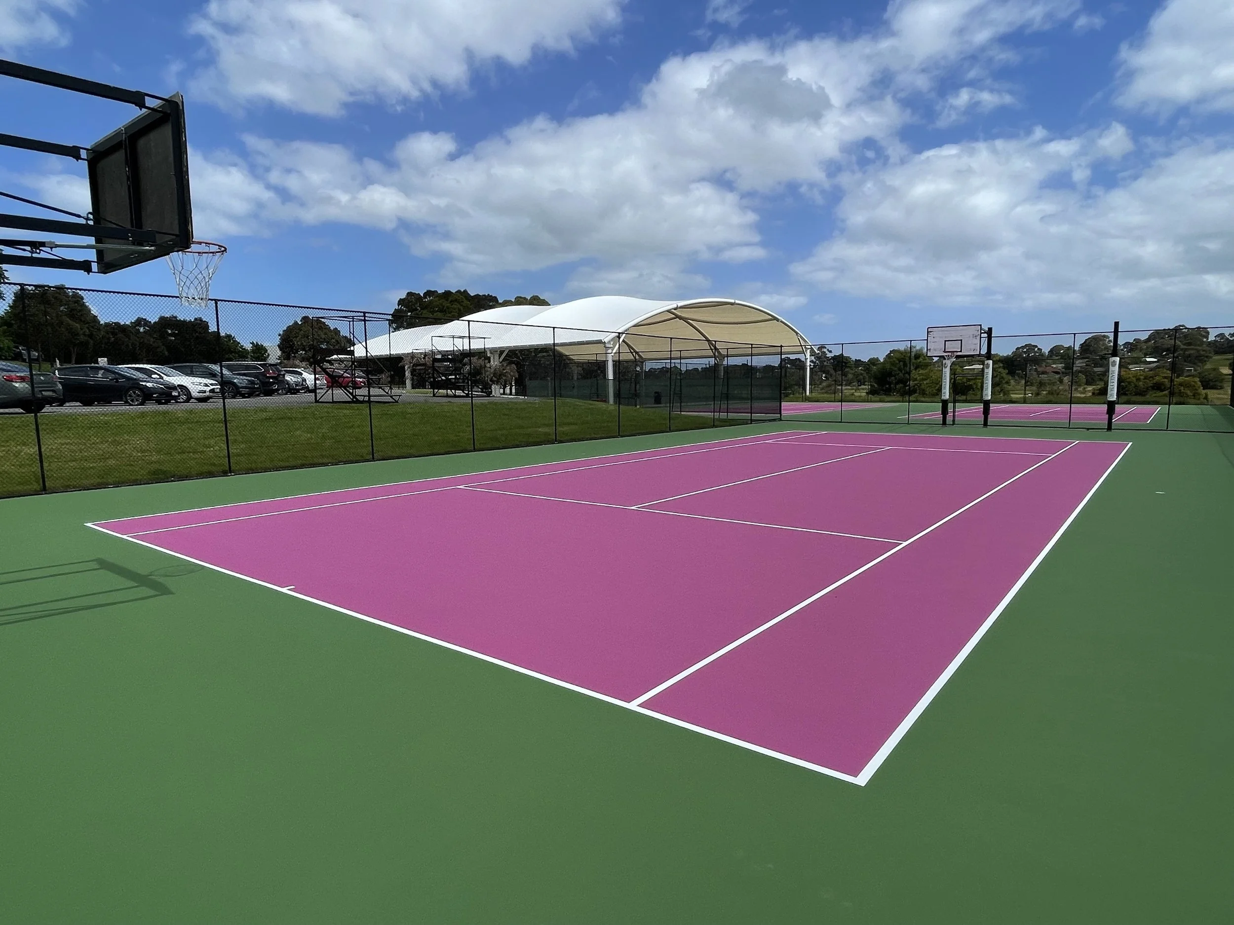 Colorful outdoor basketball and tennis courts with parking lot and trees in the background under a partly cloudy sky.