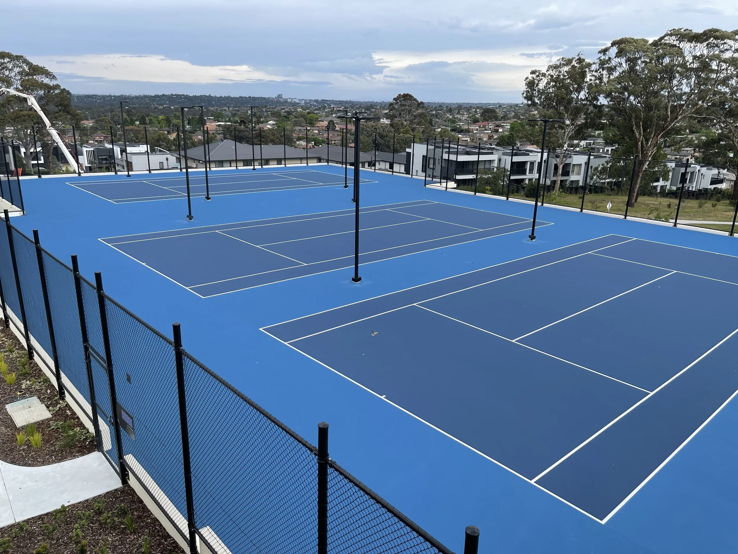 Blue-painted tennis courts on a roof, surrounded by black fencing and metal poles, with a cityscape and trees in the background under a cloudy sky.