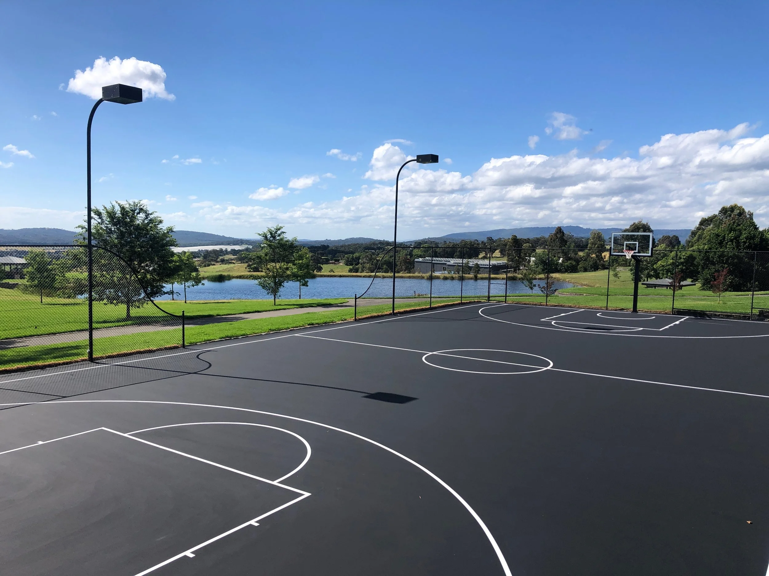 An outdoor basketball court with a black asphalt surface and white lines, surrounded by a chain-link fence, overlooking a lake and green hills under a blue sky with scattered clouds.