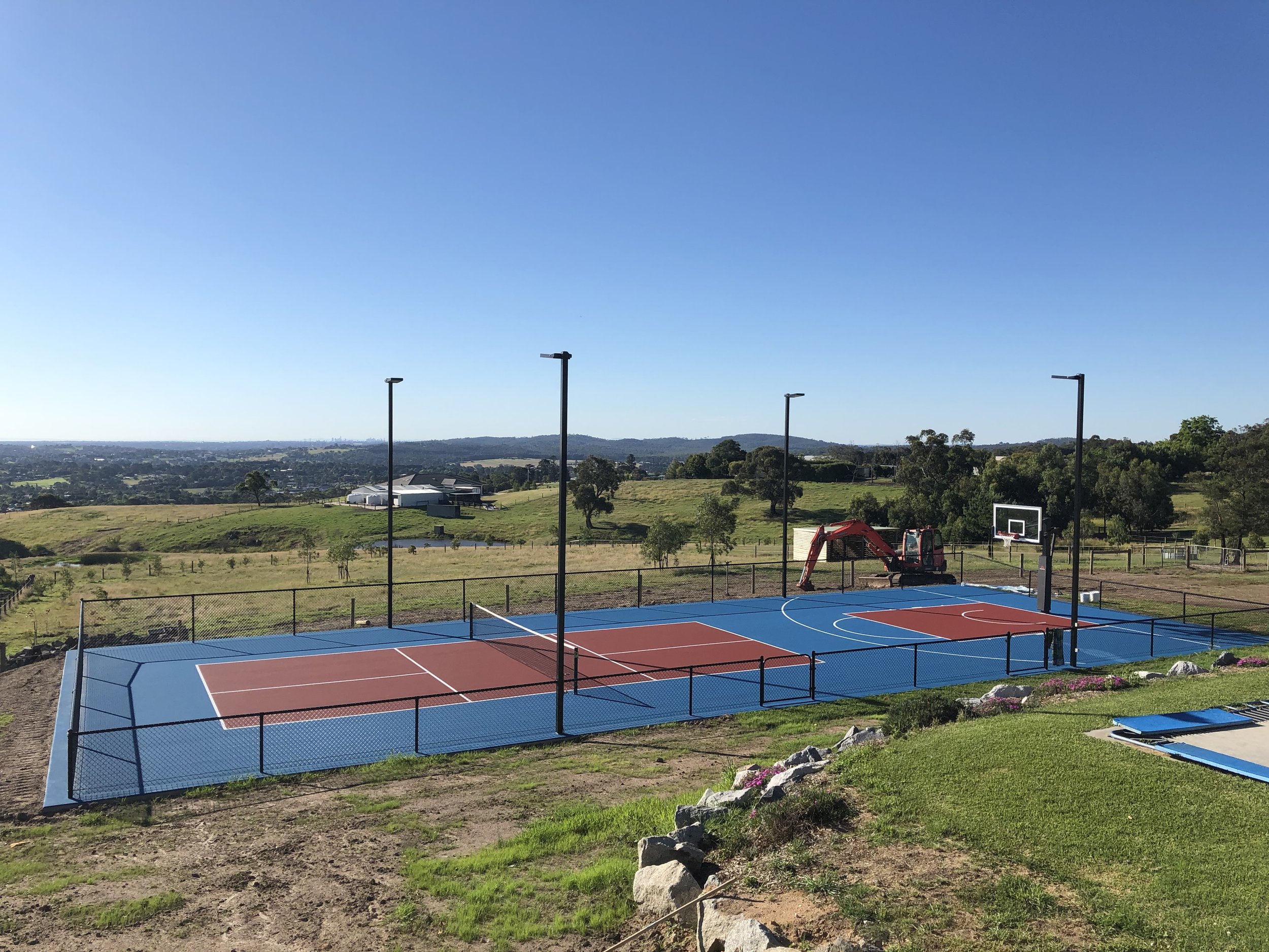 An outdoor basketball and tennis court with a black chain-link fence, surrounded by green grass and hills, under a clear blue sky.