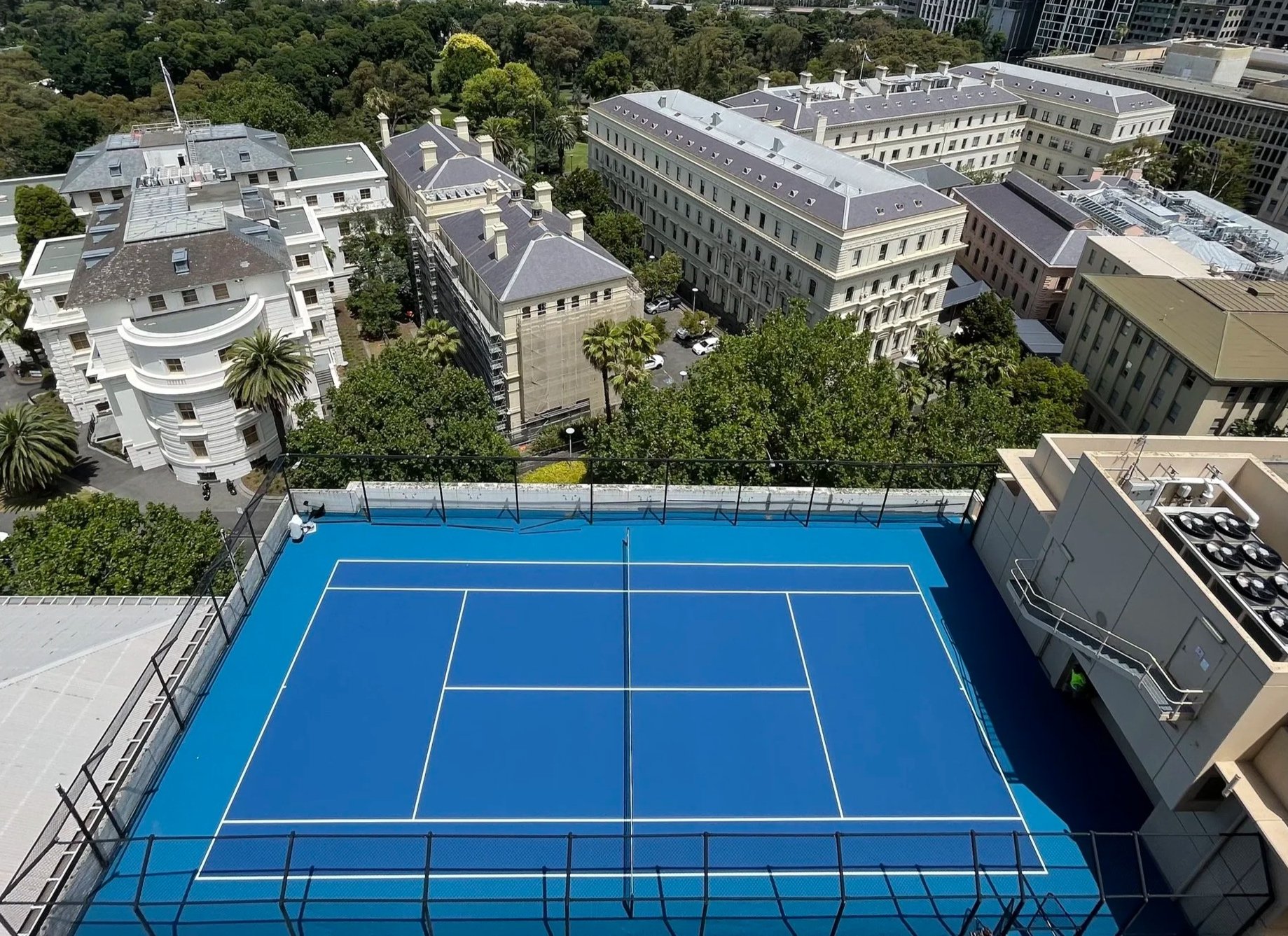 A blue rooftop tennis court with white lines and a surrounding black fence, located on top of a building in an urban area with trees and various large buildings in the background.