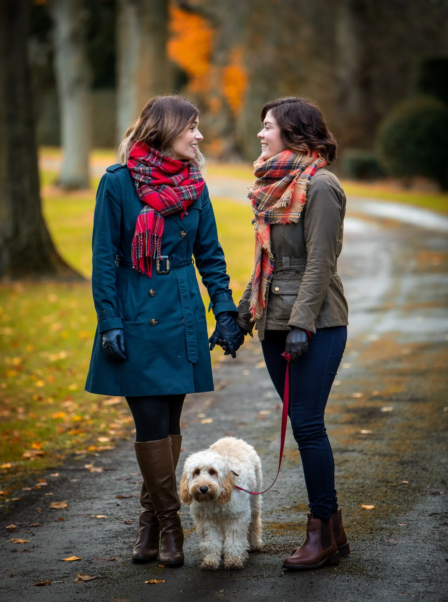 Two women standing on a park pathway, holding hands and smiling at each other, with a Golden Doodle dog standing between them on a leash. They are dressed warmly in jackets, scarves, and boots, and the background features trees with autumn foliage.