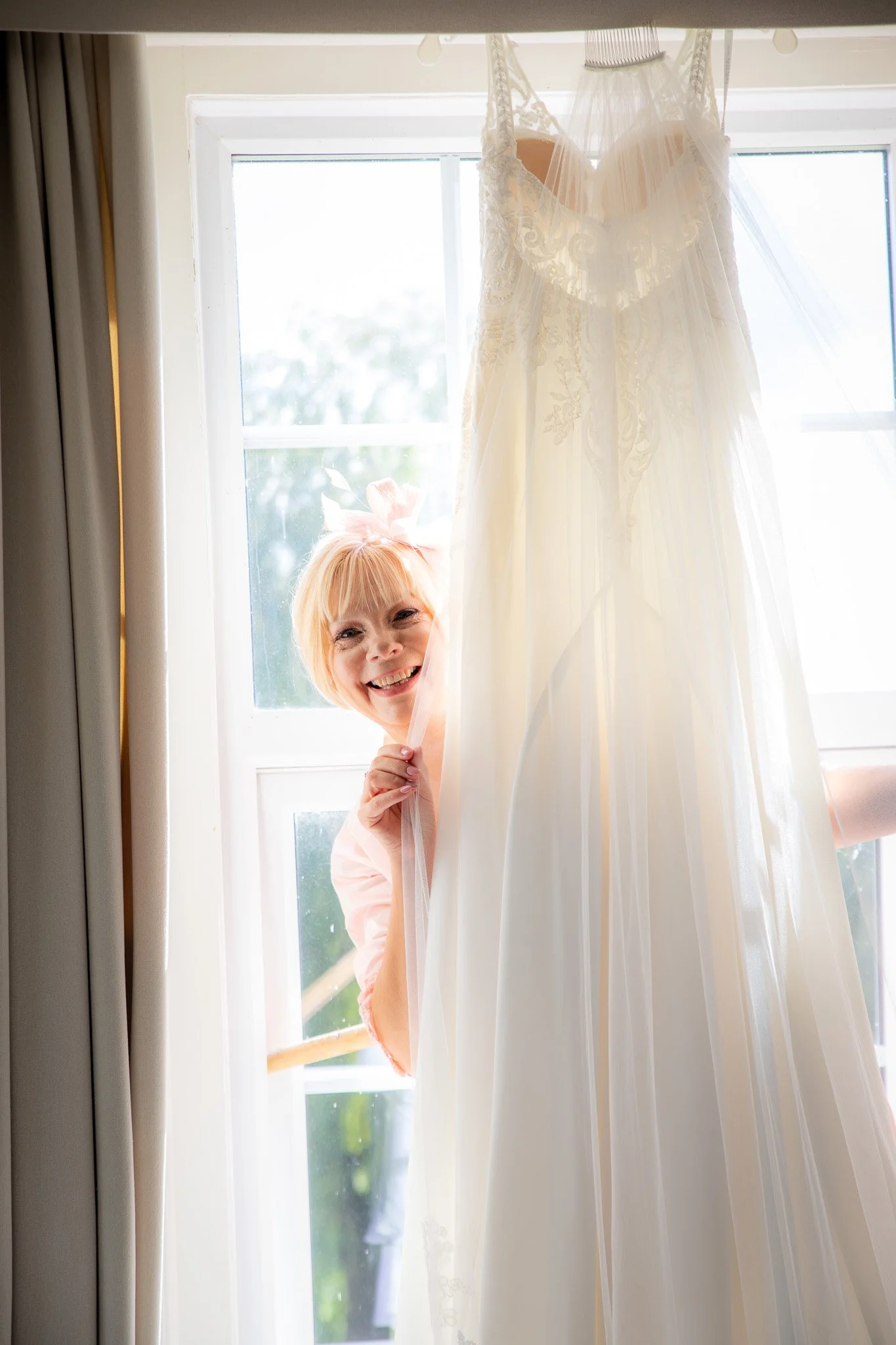 A woman with blonde hair and a pink bow headband smiling and peeking through a window behind white curtains, with a wedding dress hanging near the window.