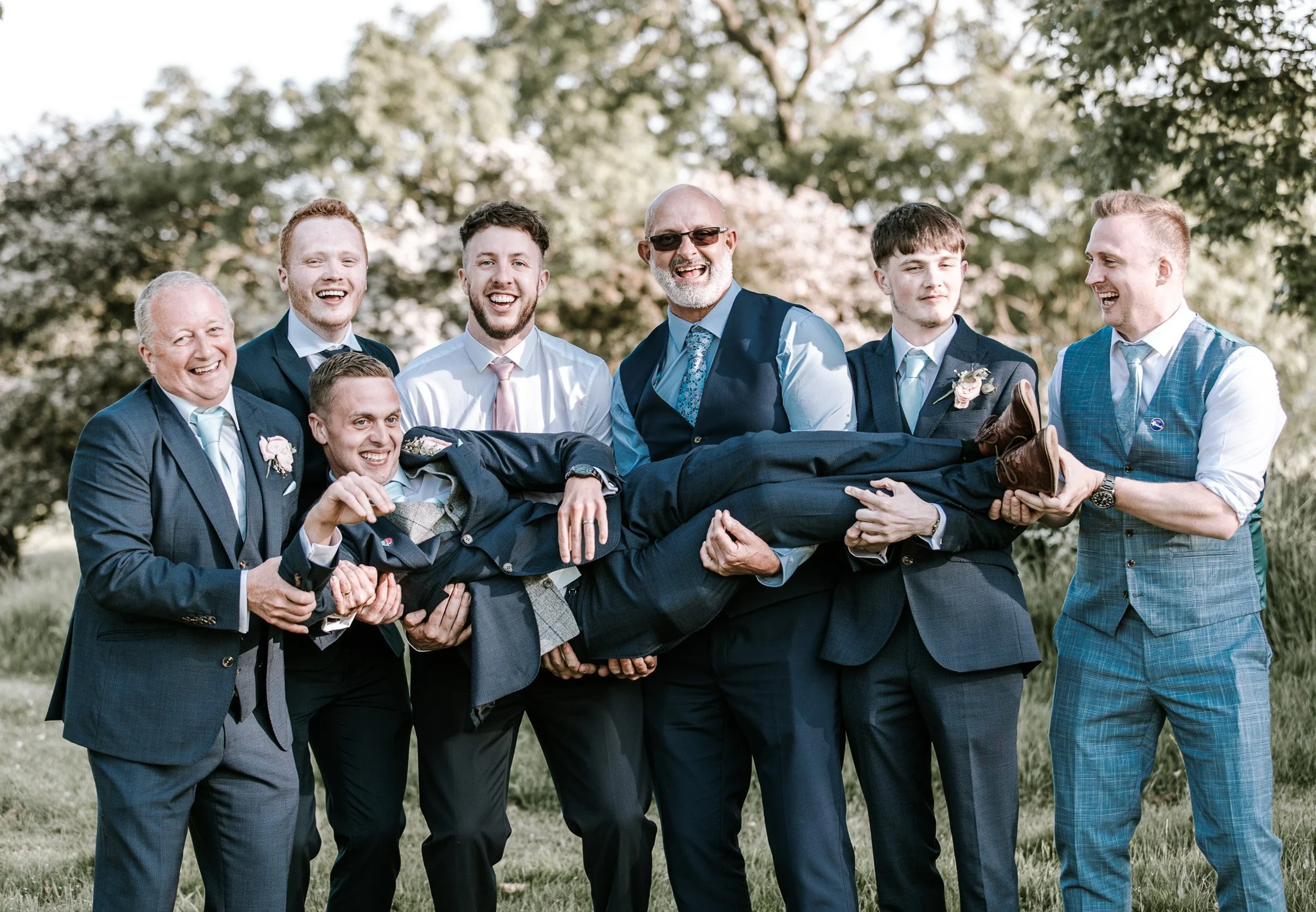 A group of men in formal suits and ties, with one man in a blue vest, are outdoors in a park-like setting, holding up a man dressed similarly in a laid-back manner. They are all smiling and enjoying each other's company.