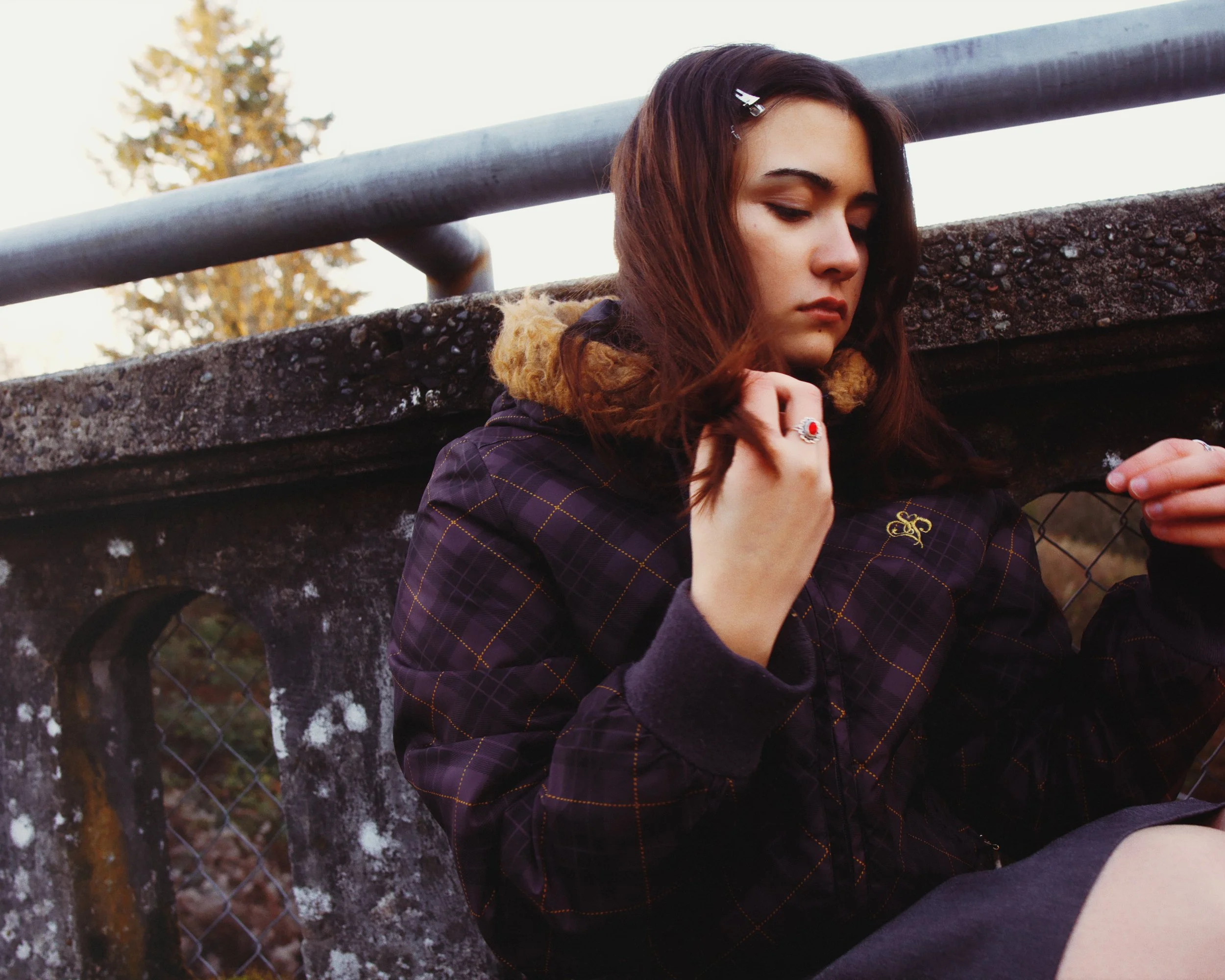 A young woman with brown hair, wearing a dark plaid jacket with a hood, sitting on a concrete bench outdoors during sunset, looking down and holding a small object or string in her hand.