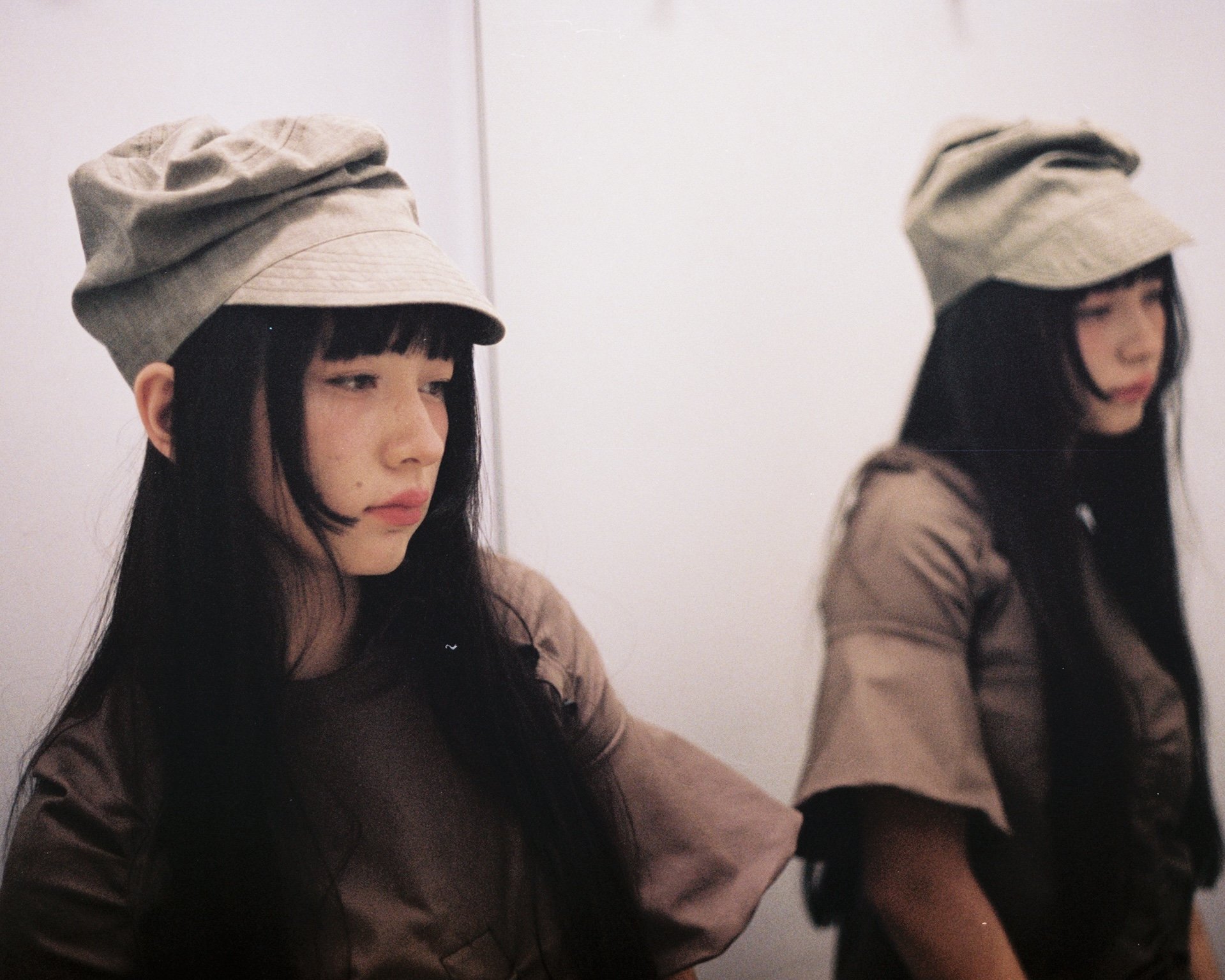 A young woman with long dark hair and bangs wearing a beige hat and a brown top standing in front of a white wall, with her reflection visible in a mirror.