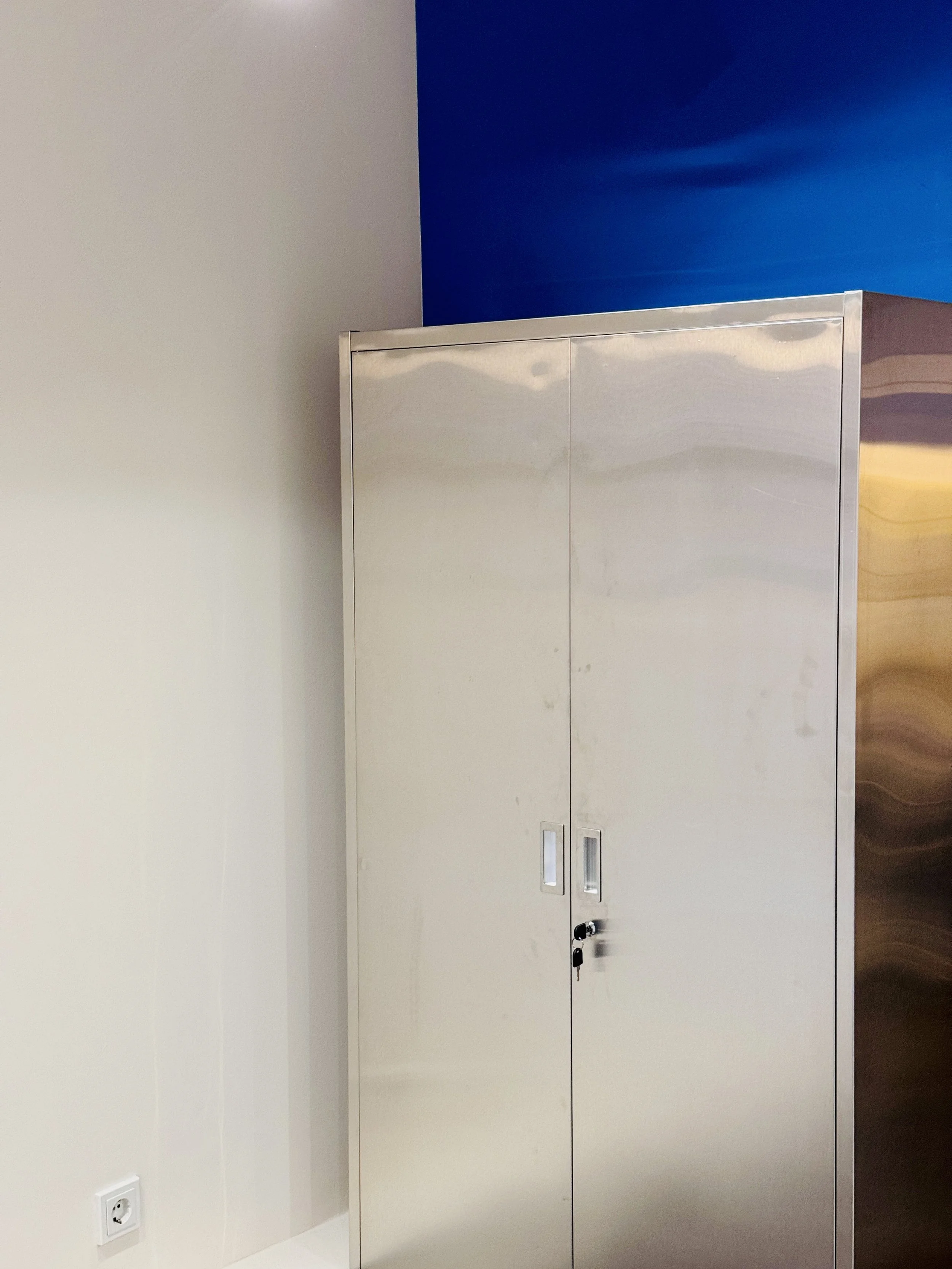 Ein rechteckiger, silberner Schrank mit doppelten Türen und Schlössern, steht vor einer weißen Wand und einer blauen Wand in einem Raum.