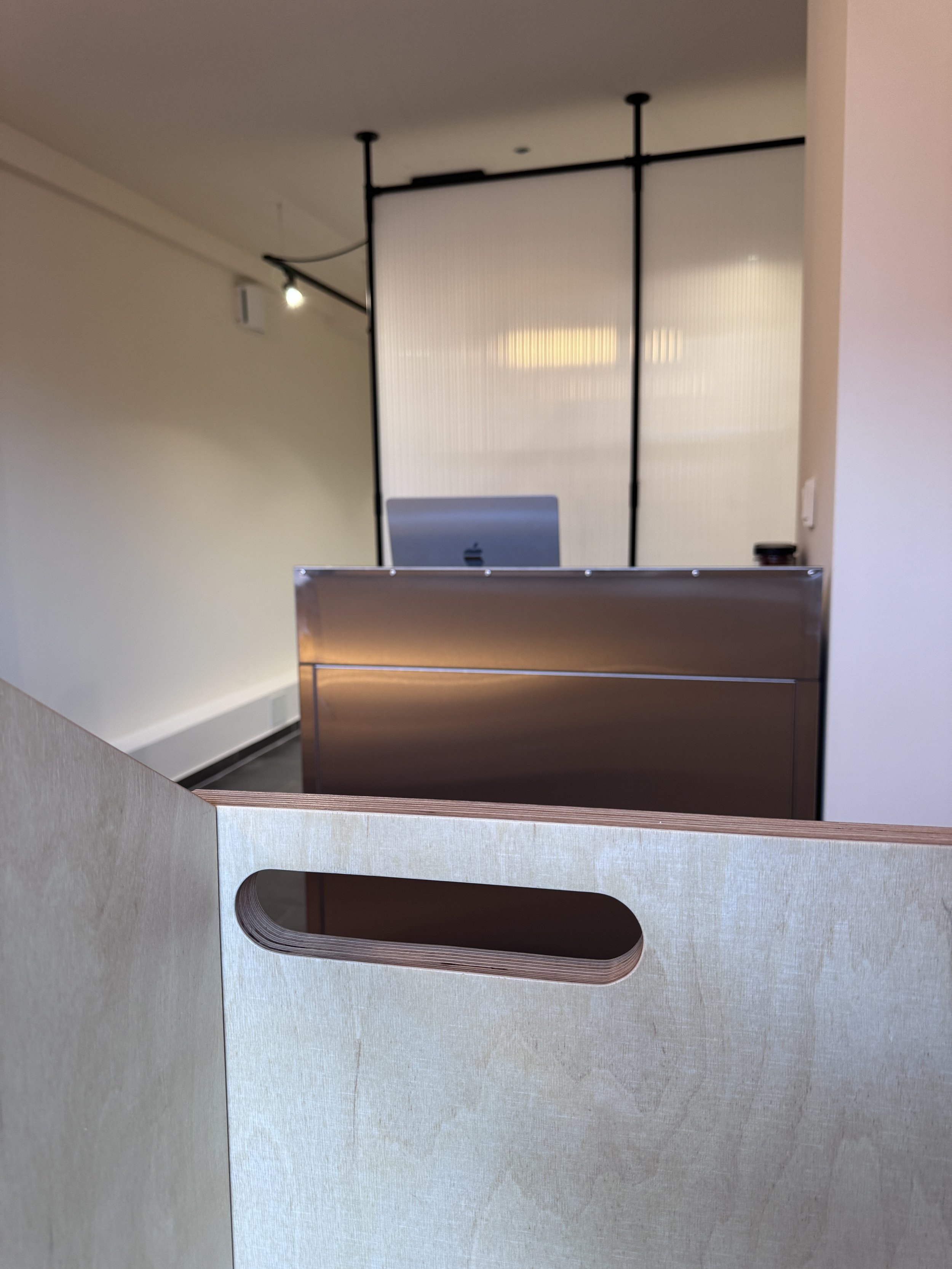 Innenaufnahme eines modernen Empfangstresens bei snyggs in Hürth-Kendenich. Wir arbeiten mit digitalen Nbuchungsmöglichkeiten. Dahinter ein halbtransparentes Raumtrennglas, im Vordergrund eine Holzwand mit Aussparung.