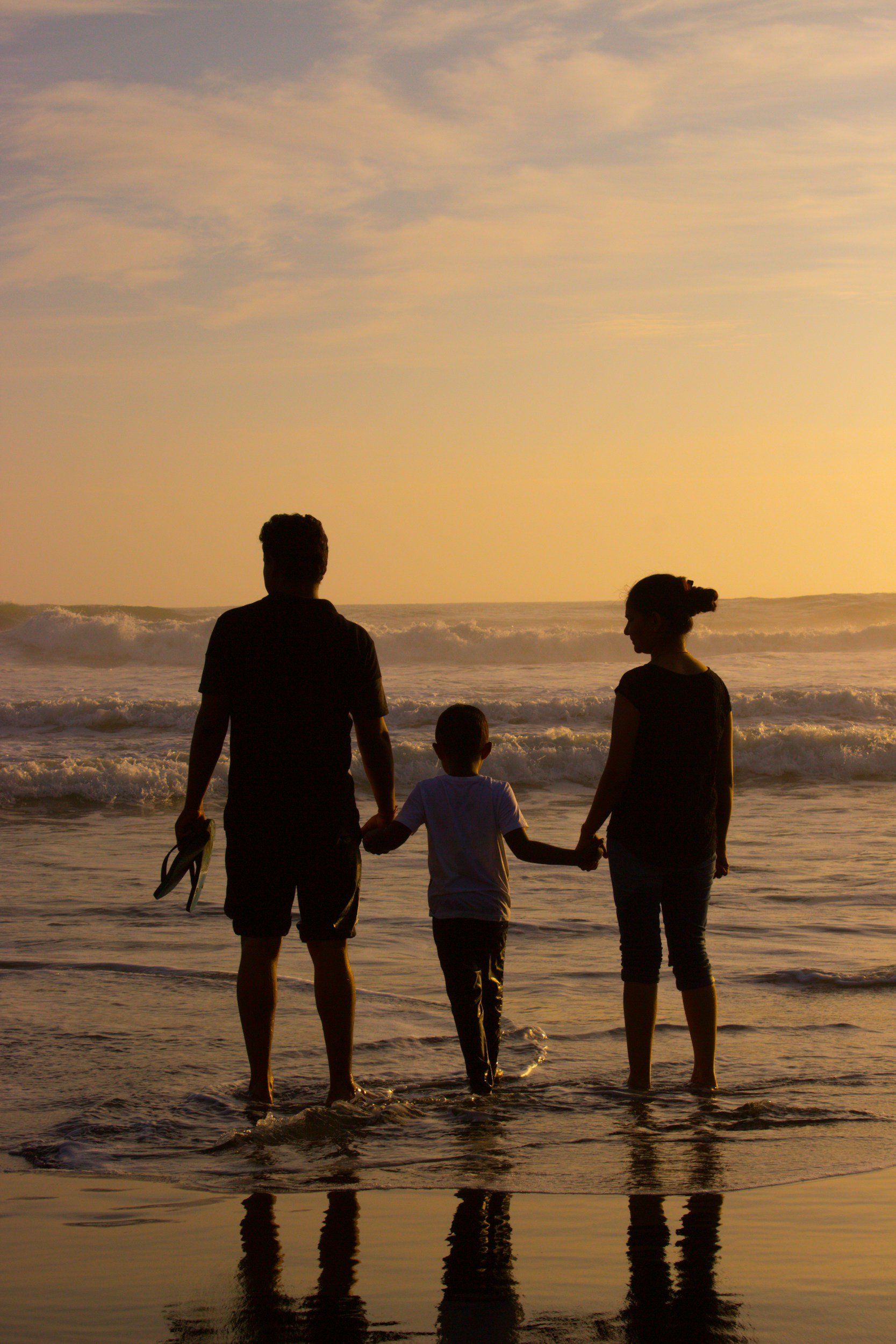 Silhouettes of a man, woman, and child holding hands at the beach during sunset, with waves in the background.