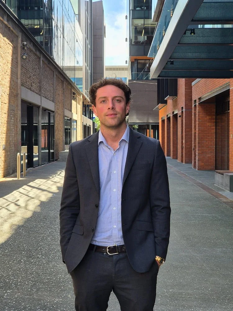 A man in a dark suit with a light blue shirt standing outdoors on a city street between modern glass and brick buildings.