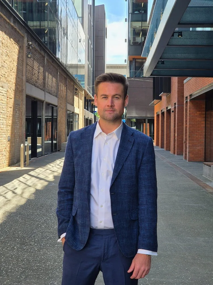 Man in a blue blazer and white shirt standing outdoors in an urban setting with modern buildings and glass facades.