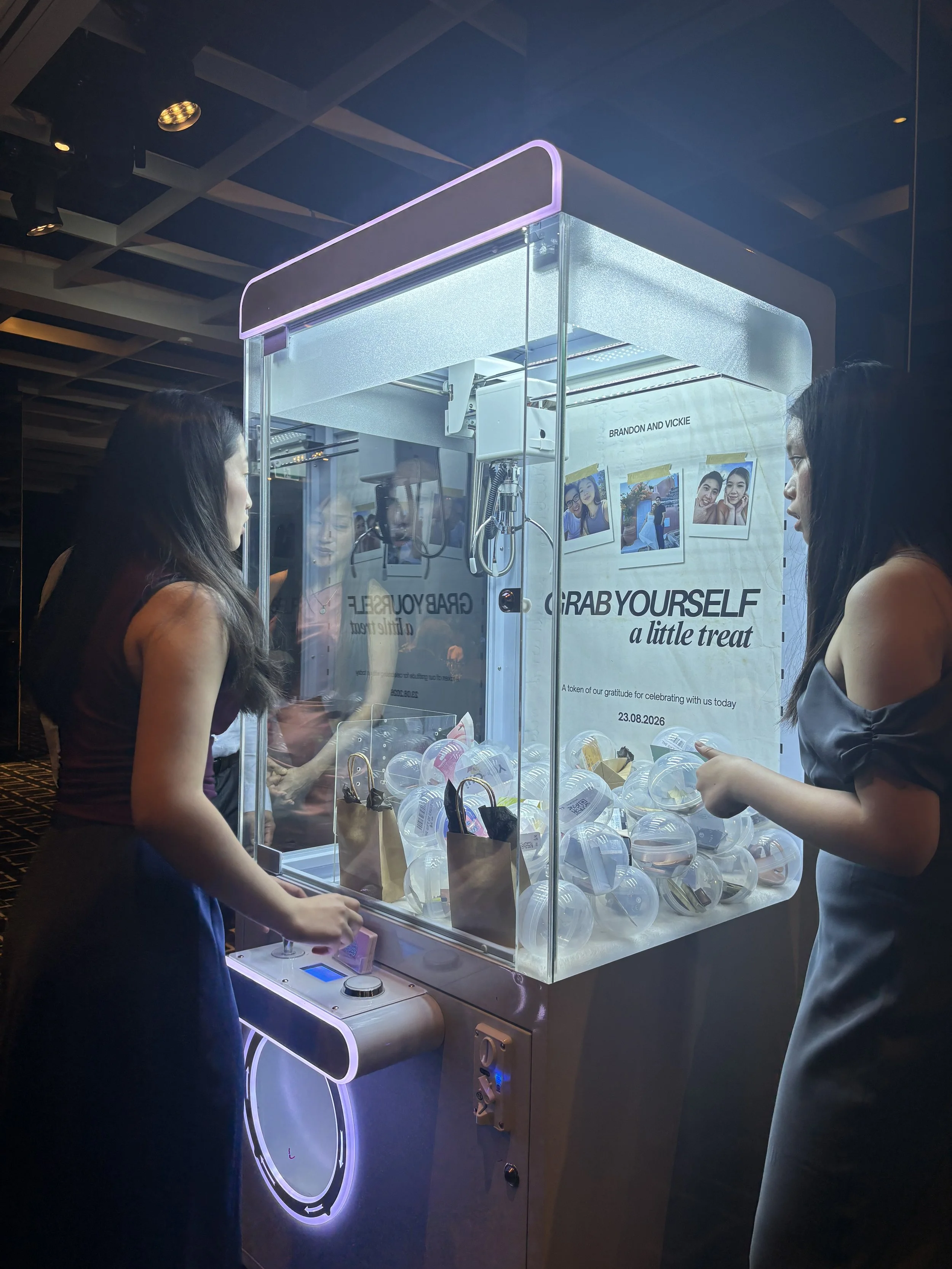 Two women playing a grab machine at an event or arcade, with a large sign behind it that reads 'Grab Yourself a Little Treat' and features photos of people and a date '23.08.2026'.