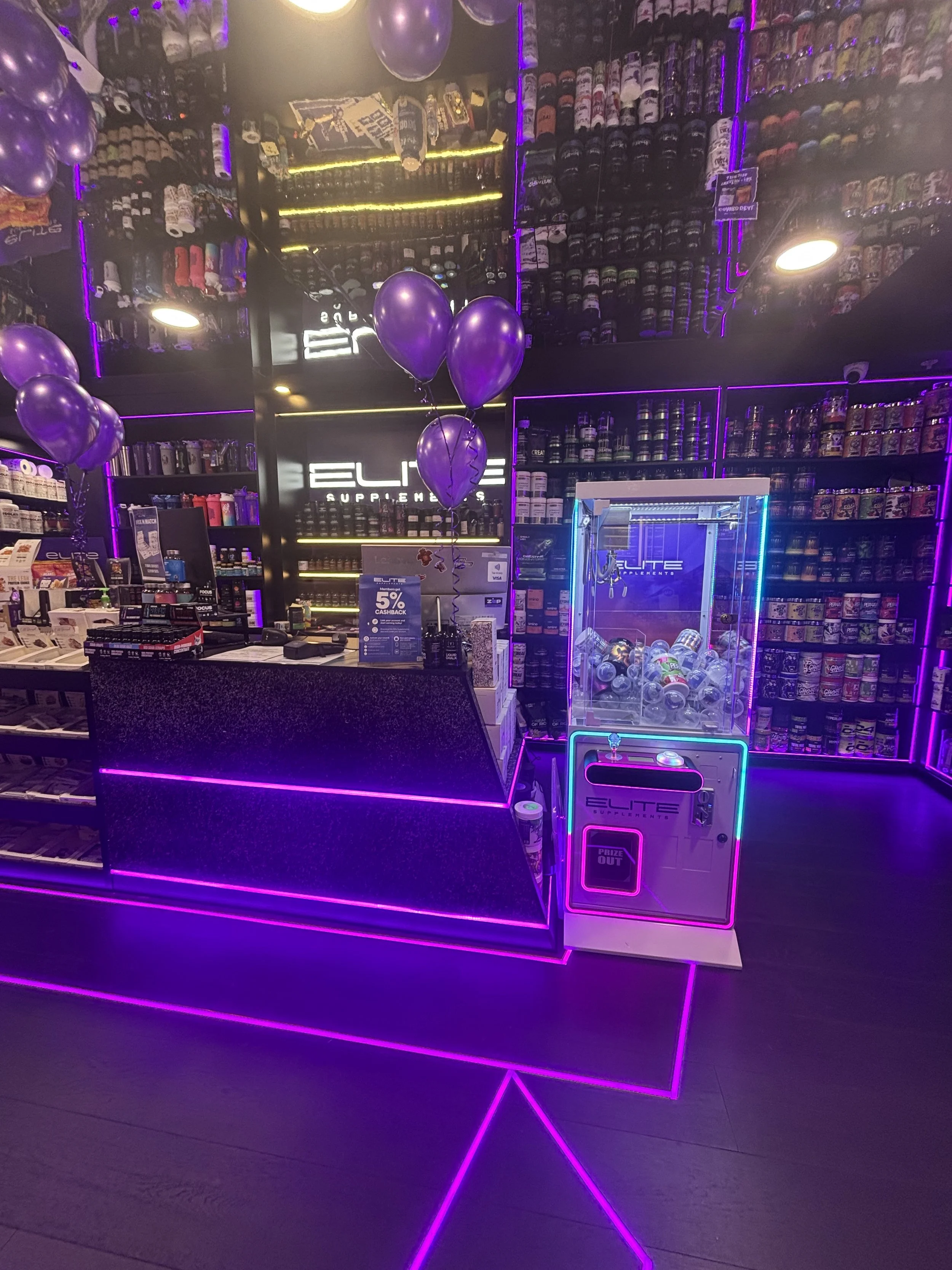 A retail store counter illuminated with purple neon lighting, displaying purple balloons, shelves stocked with supplements and other products, a claw machine filled with small plush toys, and a sign promoting a cashback offer.