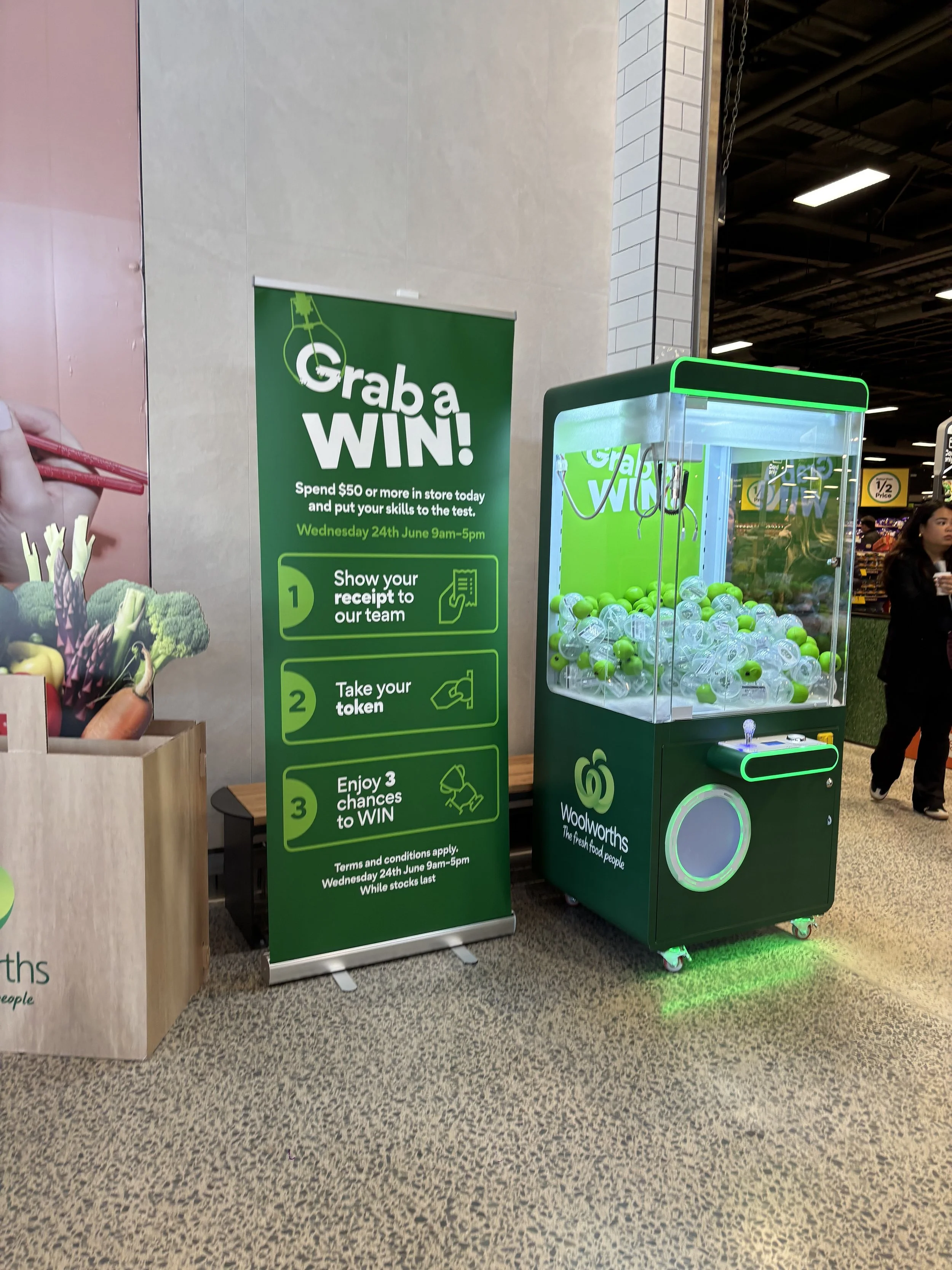 A promotional display at Woolworths supermarket with a green sign advertising a win contest for spending $50 or more, and a green and white claw machine game filled with green and clear plastic balls.