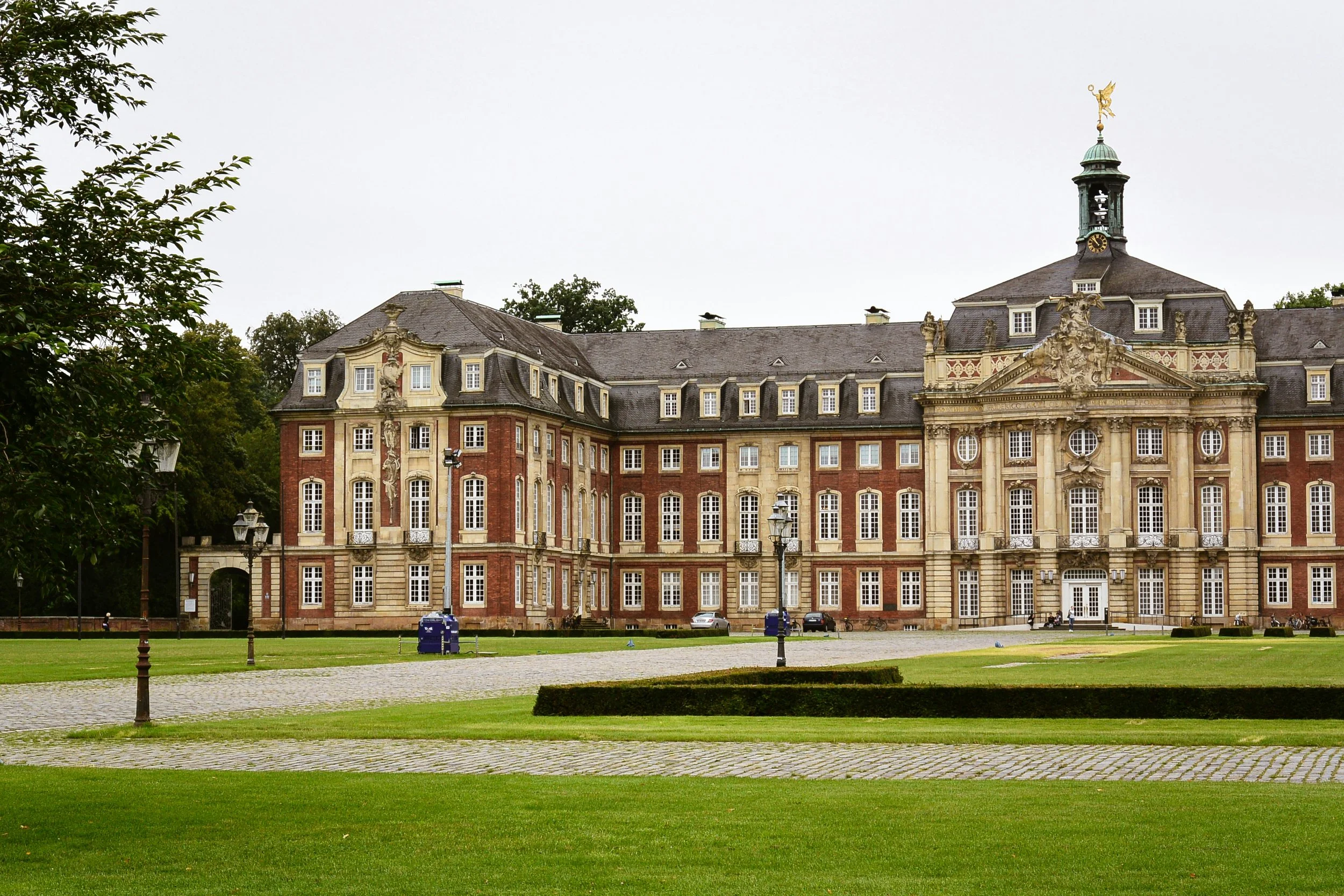 A historic European-style palace with a symmetrical facade, multiple windows, ornate decorations, and a clock tower topped with a golden figure, surrounded by a well-manicured lawn and walkway.