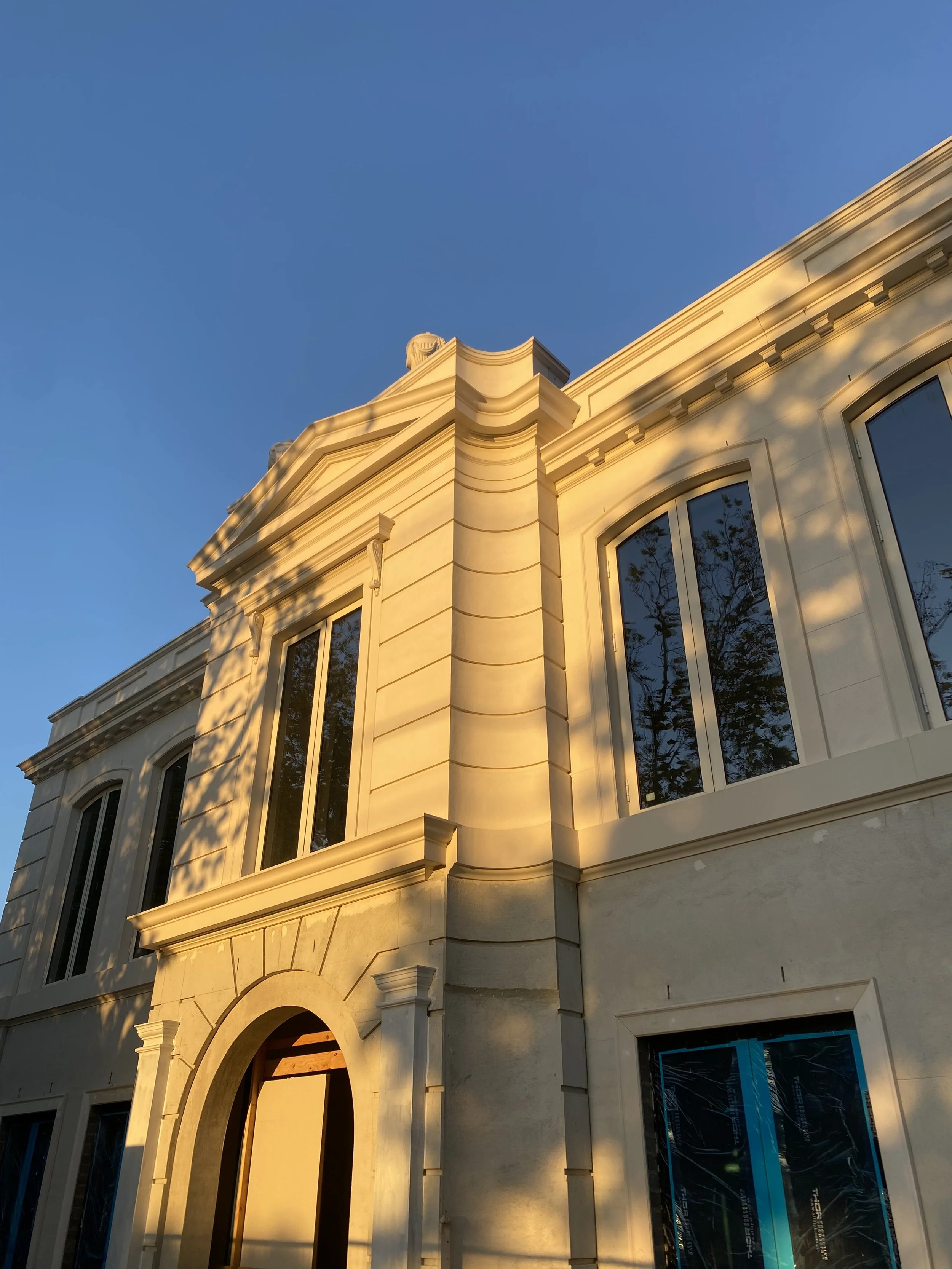 Corner of a white classical style building with large arched windows, decorative molding, and detailed cornices, illuminated by sunlight against a clear blue sky.