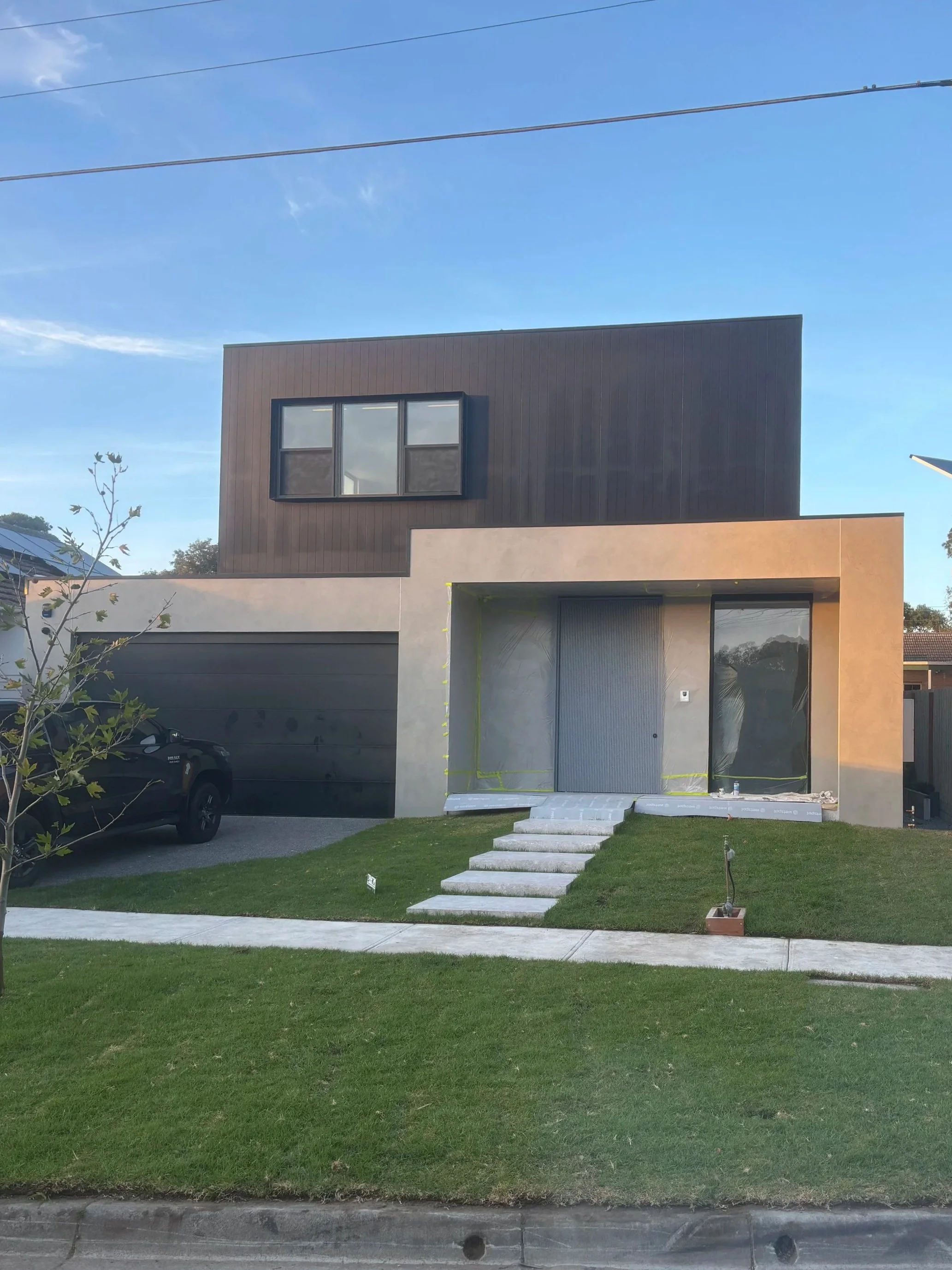 Modern, two-story house with dark brown upper level, beige lower level, large window on the front, and a small front yard with a green lawn and white pathway stairs.