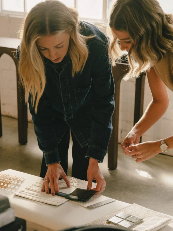 Deux femmes regardent des échantillons de matériaux sur une table, dans un intérieur lumineux.